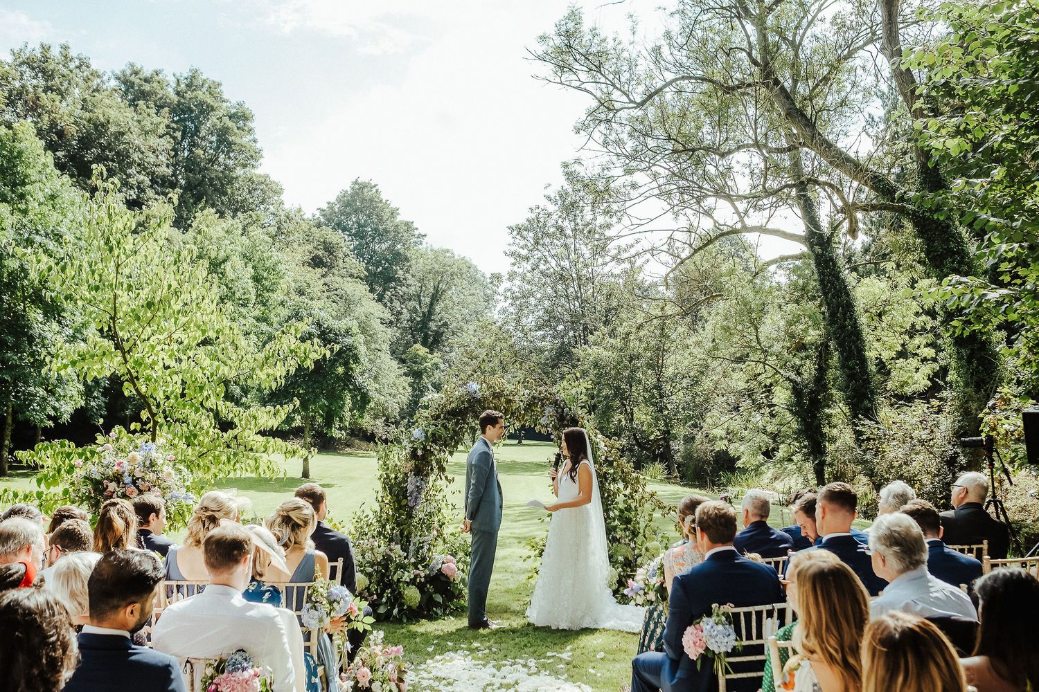 Wedding ceremony takes place under floral arch with guests seated in garden setting.