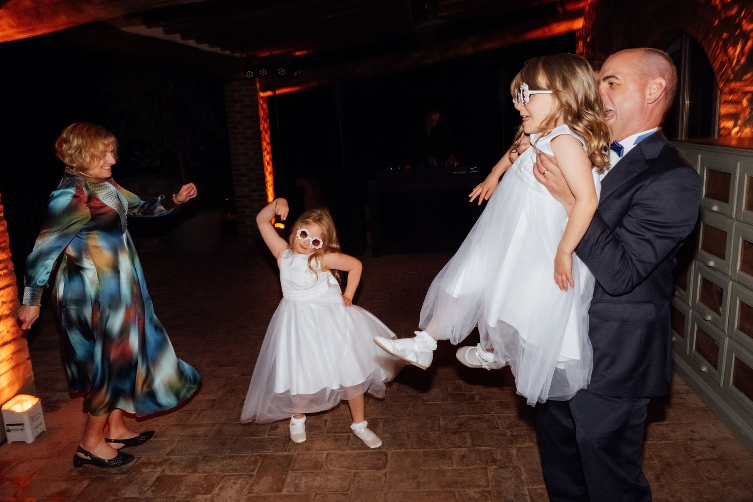 Two little girls in white dresses dance at a wedding reception while a man in a suit lifts one and a woman in a colorful dress joins in.