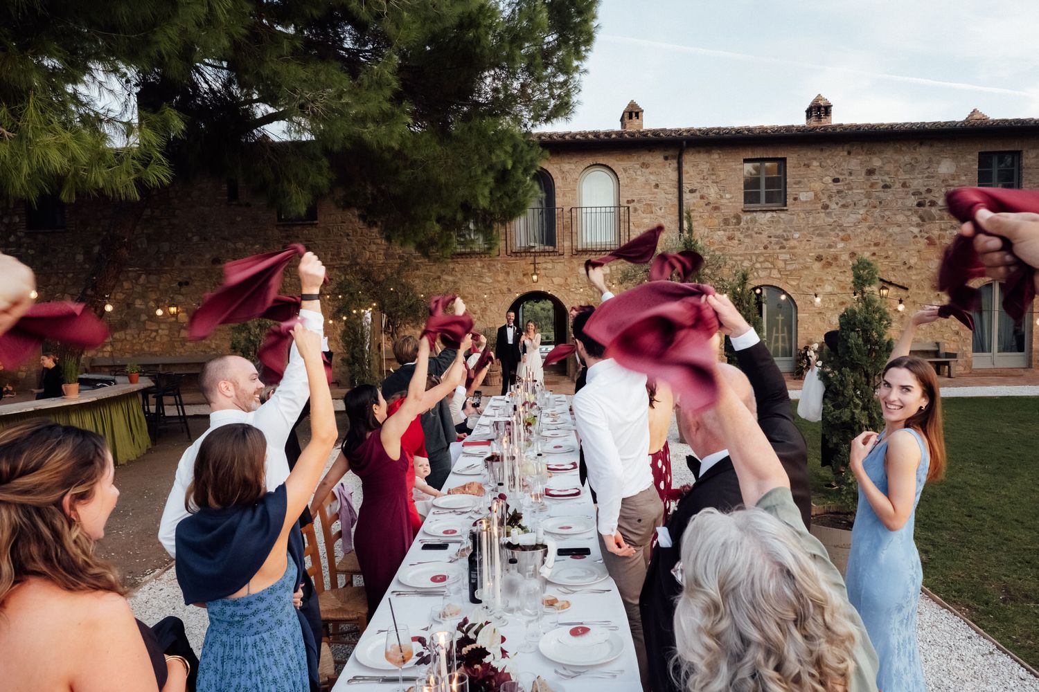 Wedding guests wave red napkins in the air at a long outdoor dinner table beside a rustic stone villa surrounded by trees.