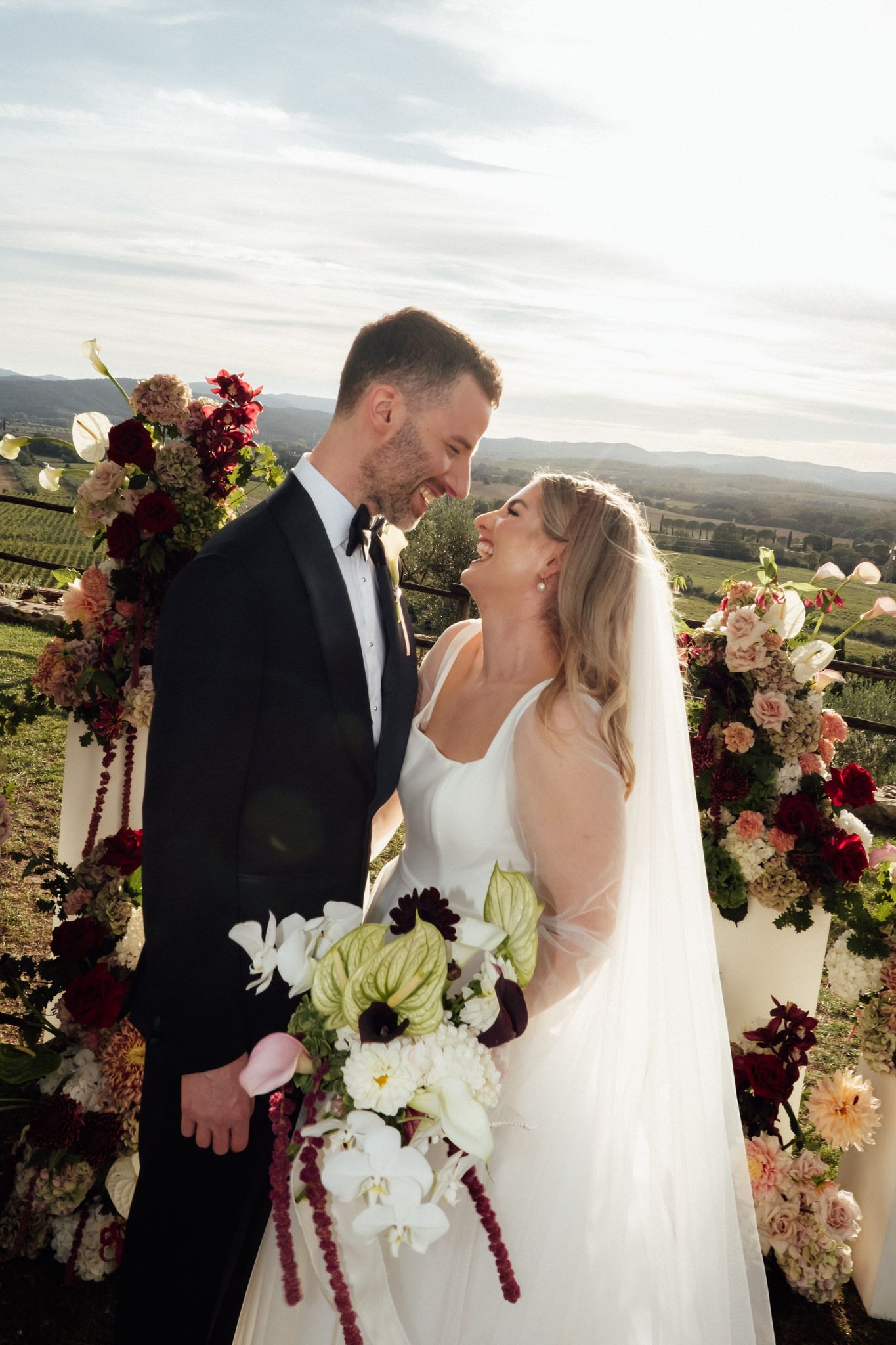 Bride and groom laugh together at a vineyard wedding ceremony, surrounded by burgundy and blush floral arrangements and a lush bouquet.