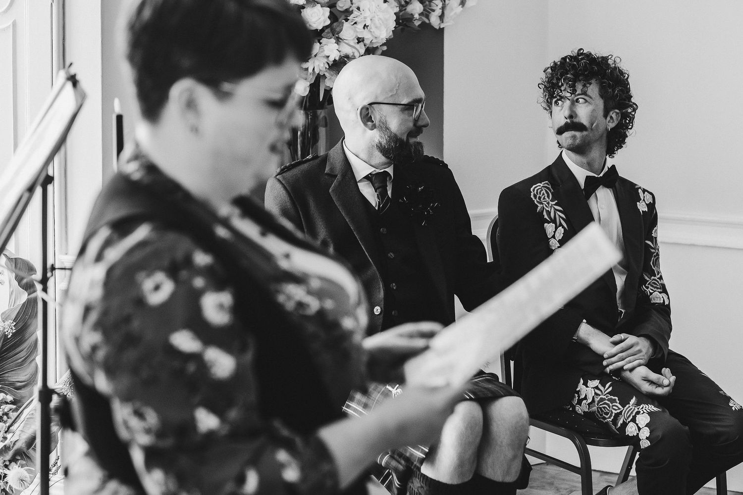 Black and white photo of a woman in a floral top reading aloud to two seated men in formal attire at an intimate wedding ceremony.