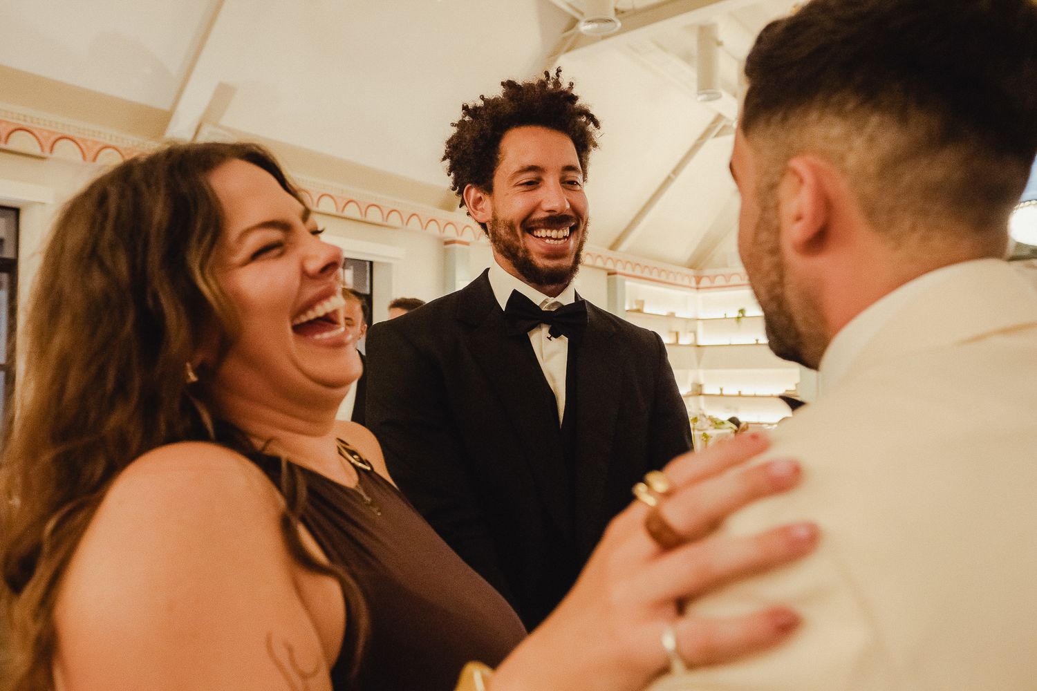 Wedding guests share joyful laughter and embrace during a reception celebration under a white tent.