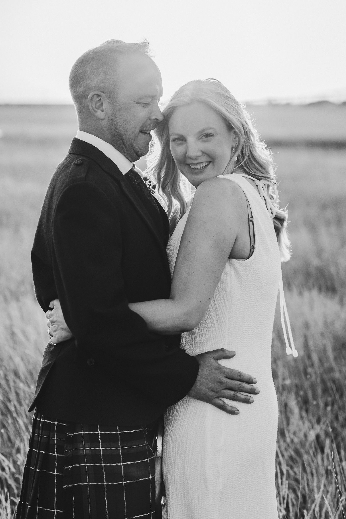 A romantic black and white portrait of a couple embracing in a field wearing wedding attire.
