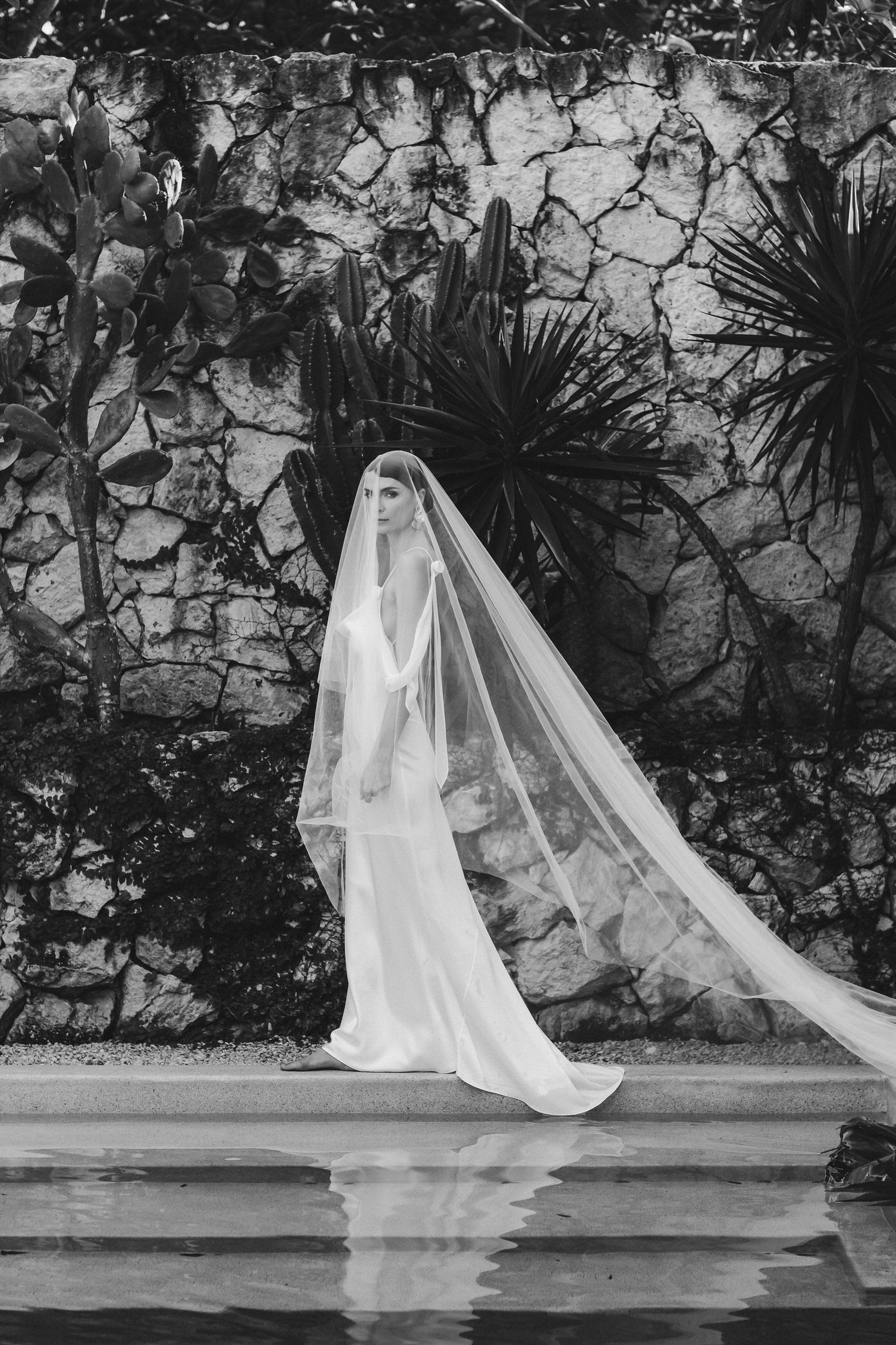 A flowing white bridal gown with long veil creates an ethereal silhouette against a stone wall and garden backdrop.