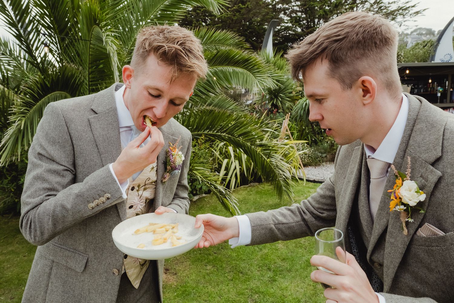 A group of groomsmen in gray suits share a playful moment eating soup together outdoors.
