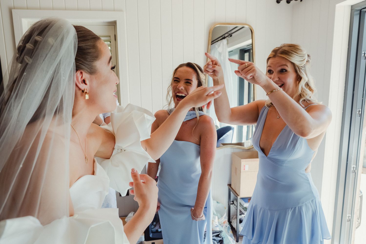 Wedding party members in light blue dresses share joyful moments while getting ready in a bright room.
