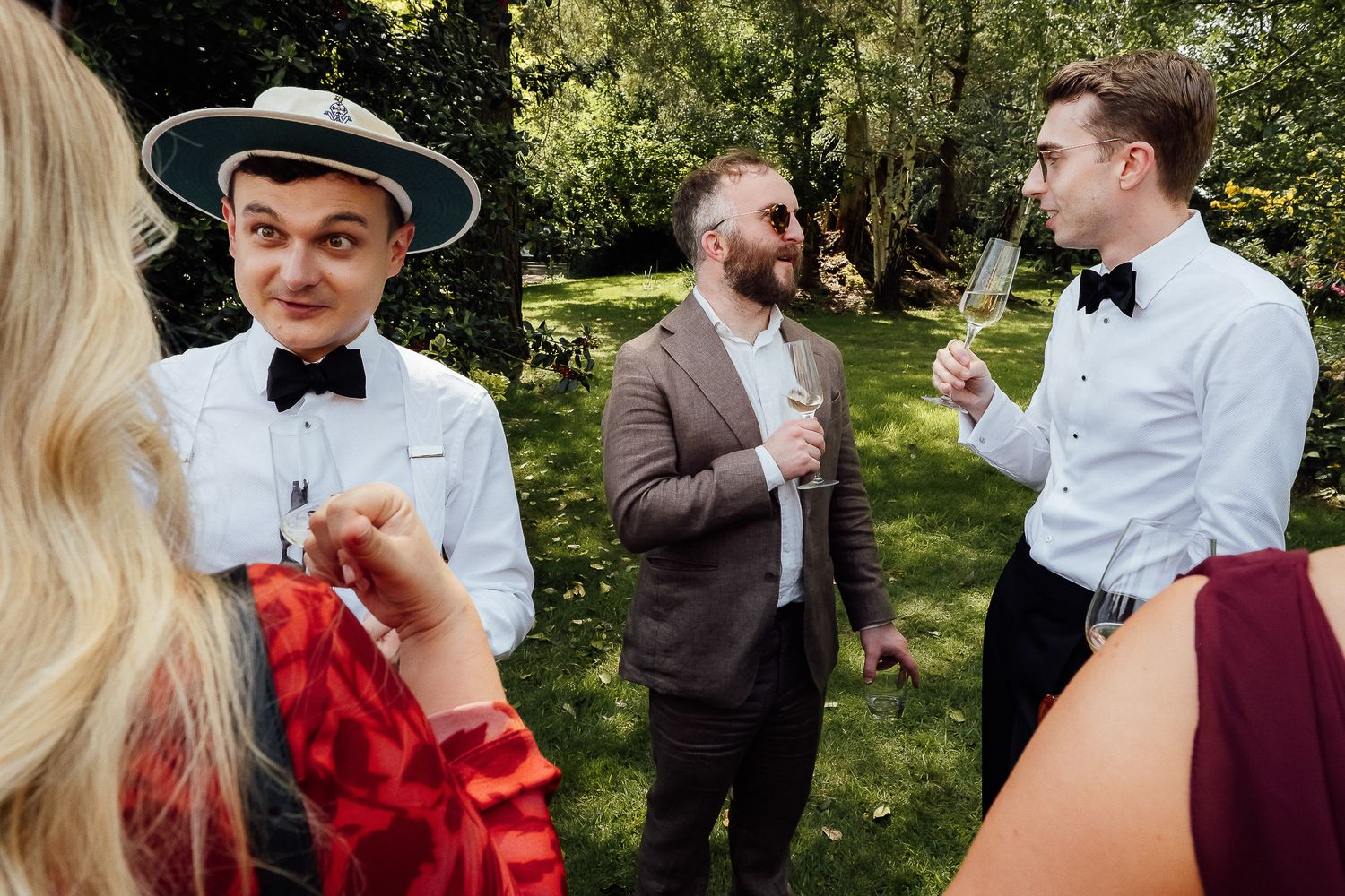 Men in formal attire with bow ties socializing outdoors at a garden wedding reception.