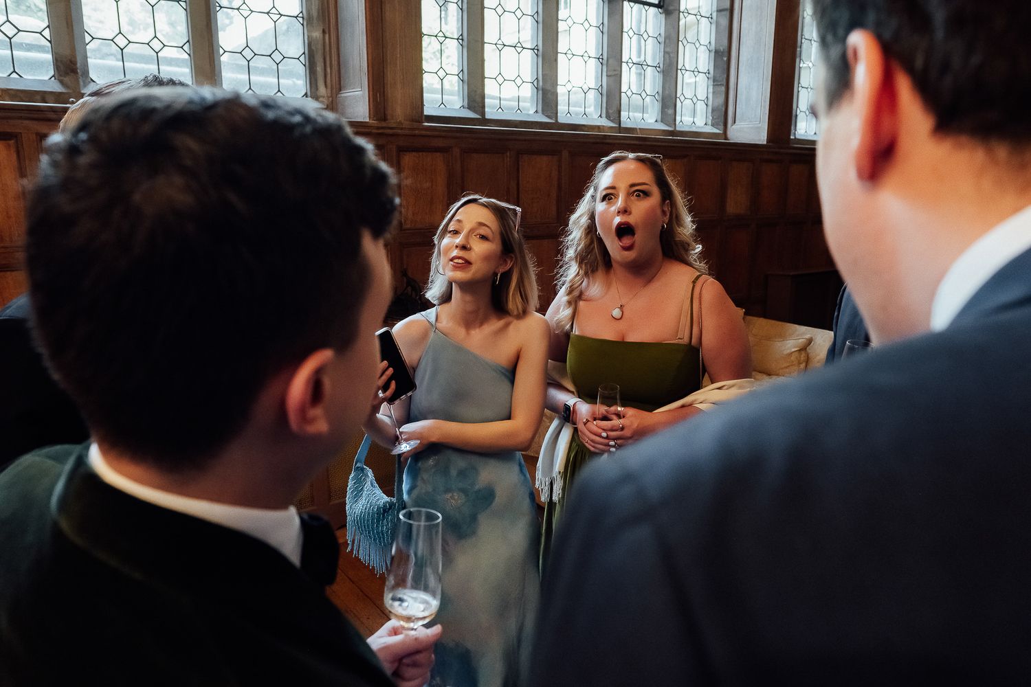 Two women in elegant dresses react with surprise and excitement while socializing with guests at a formal indoor event.