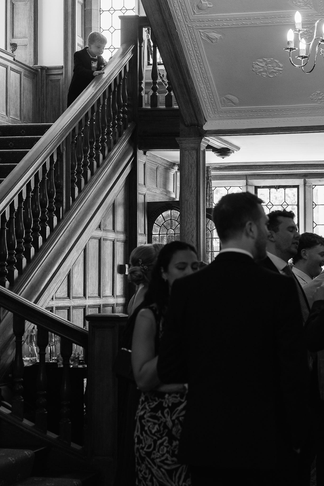 Black and white photo of elegantly dressed guests socializing near a grand wooden staircase in a historic venue.