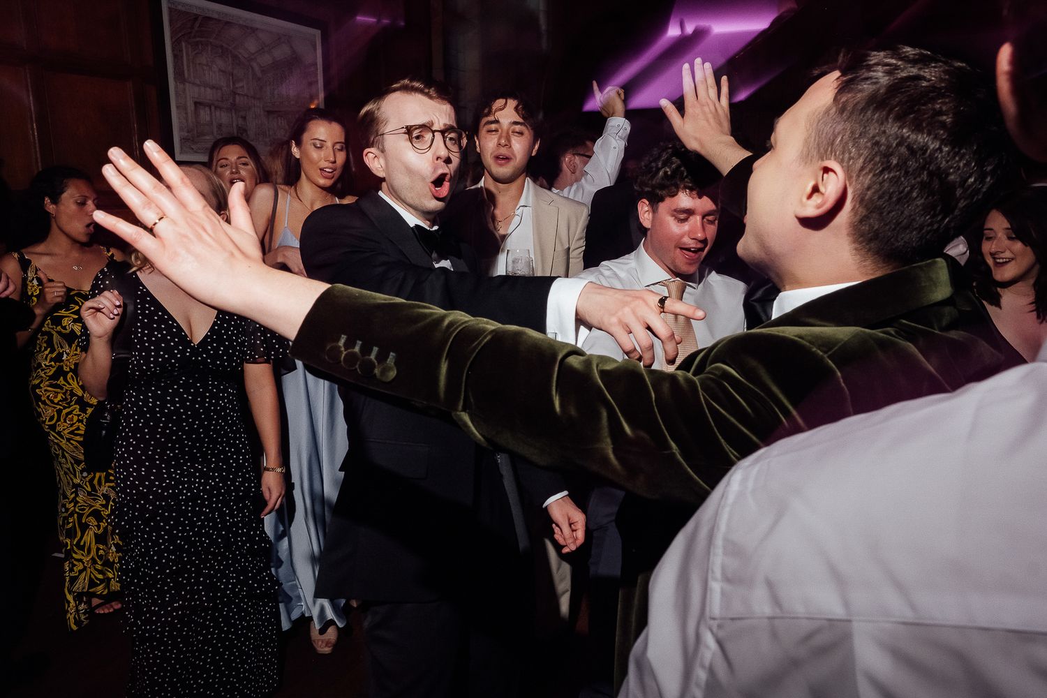 Guests dancing energetically at a wedding reception party under purple lights.