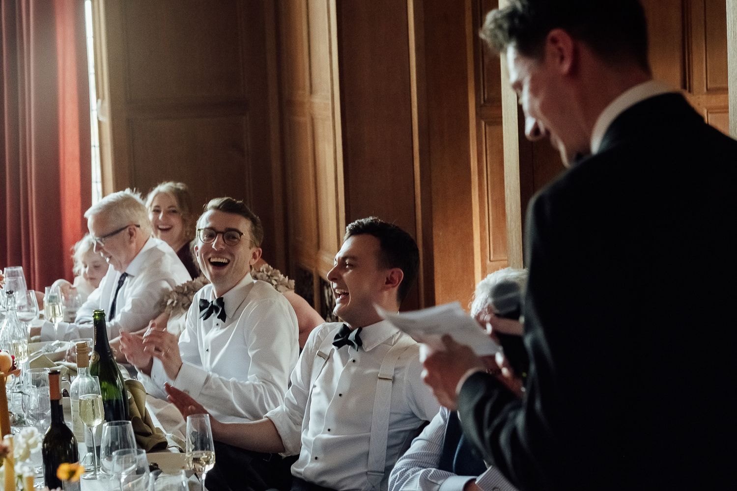 Wedding guests laughing during a humorous best man speech at an elegant formal reception dinner.