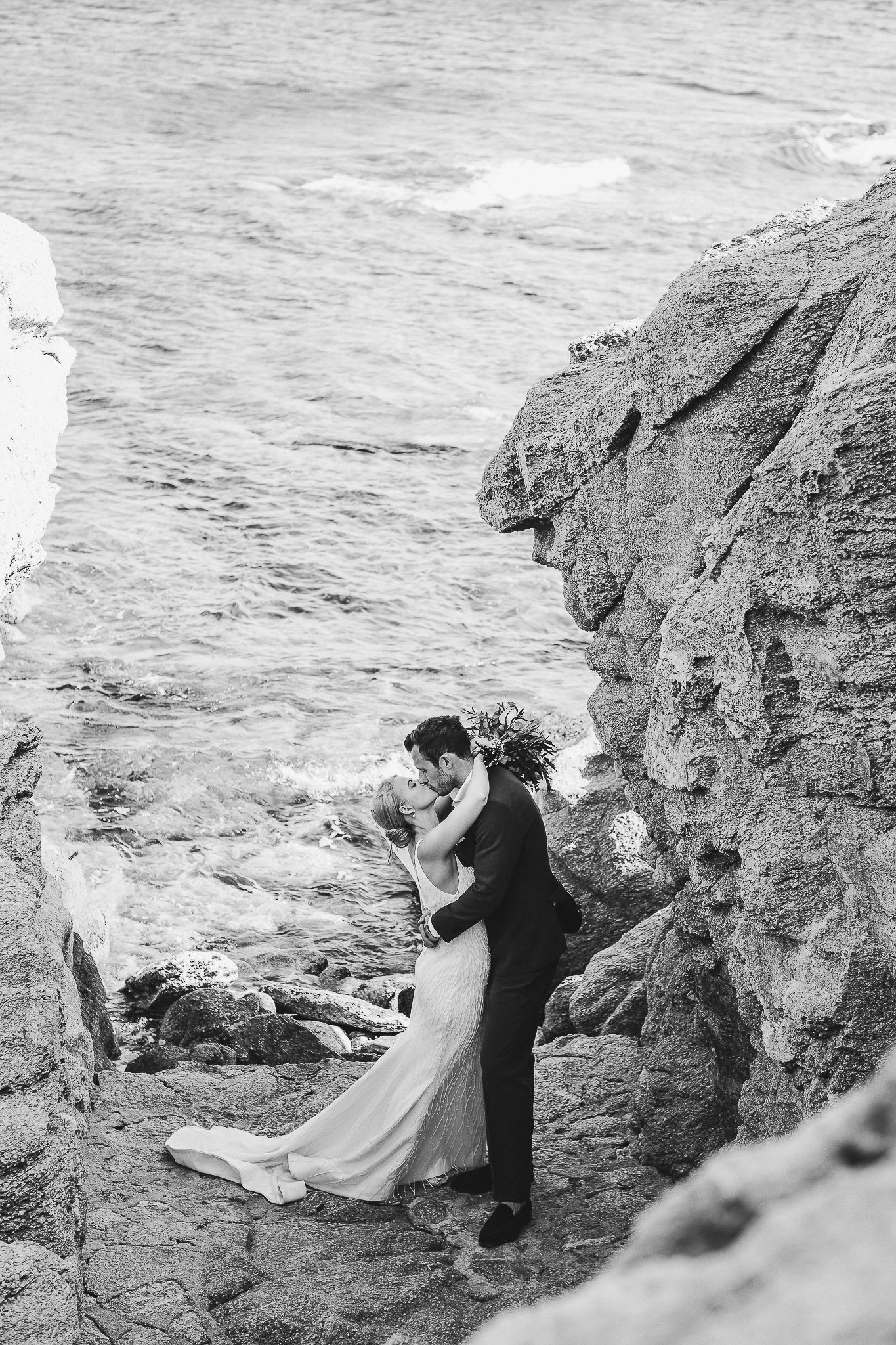 A romantic couple shares an intimate moment on rocky coastal cliffs in elegant black and white photography.