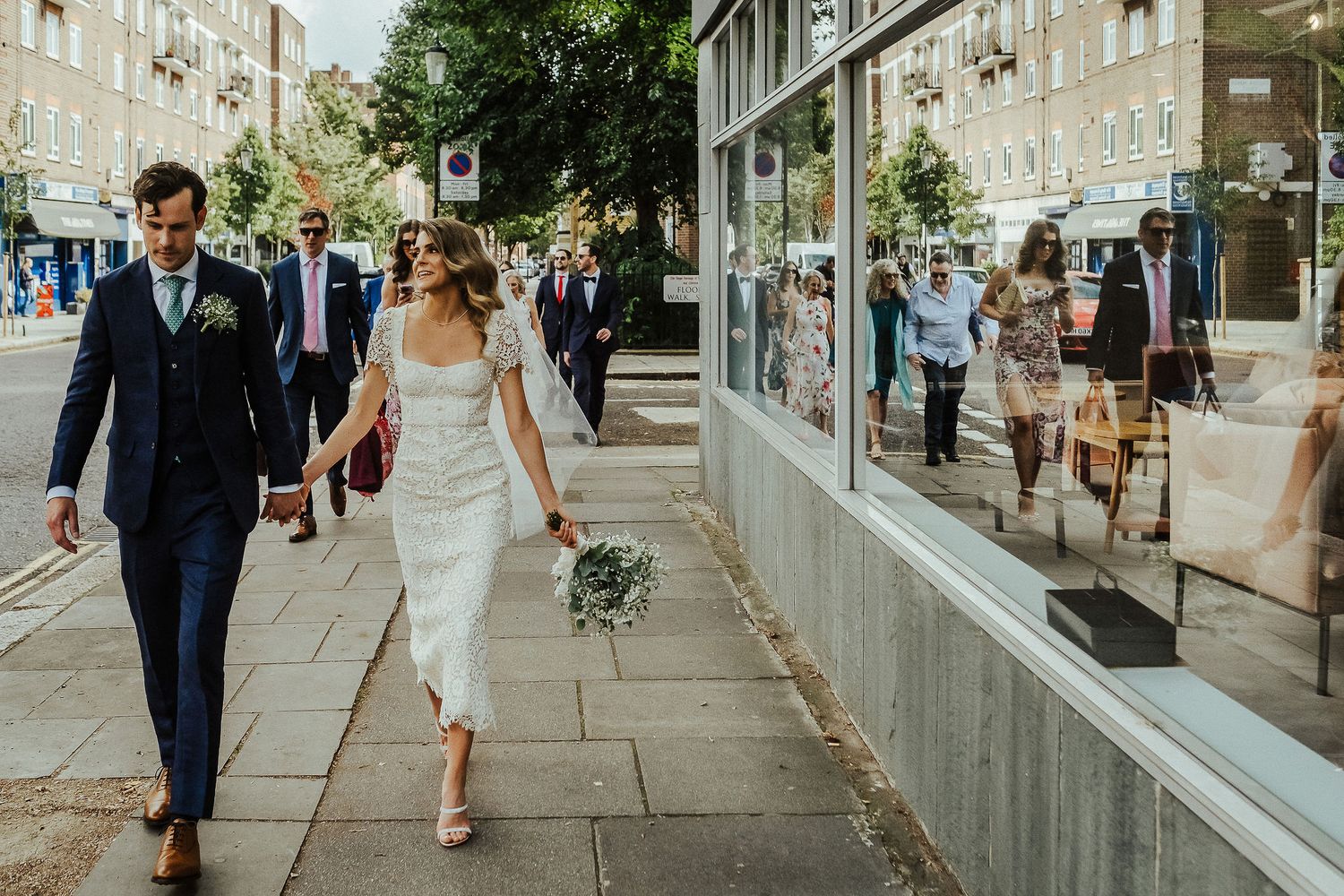 Newlyweds walk hand in hand down a sunny urban sidewalk with stone buildings in the background.