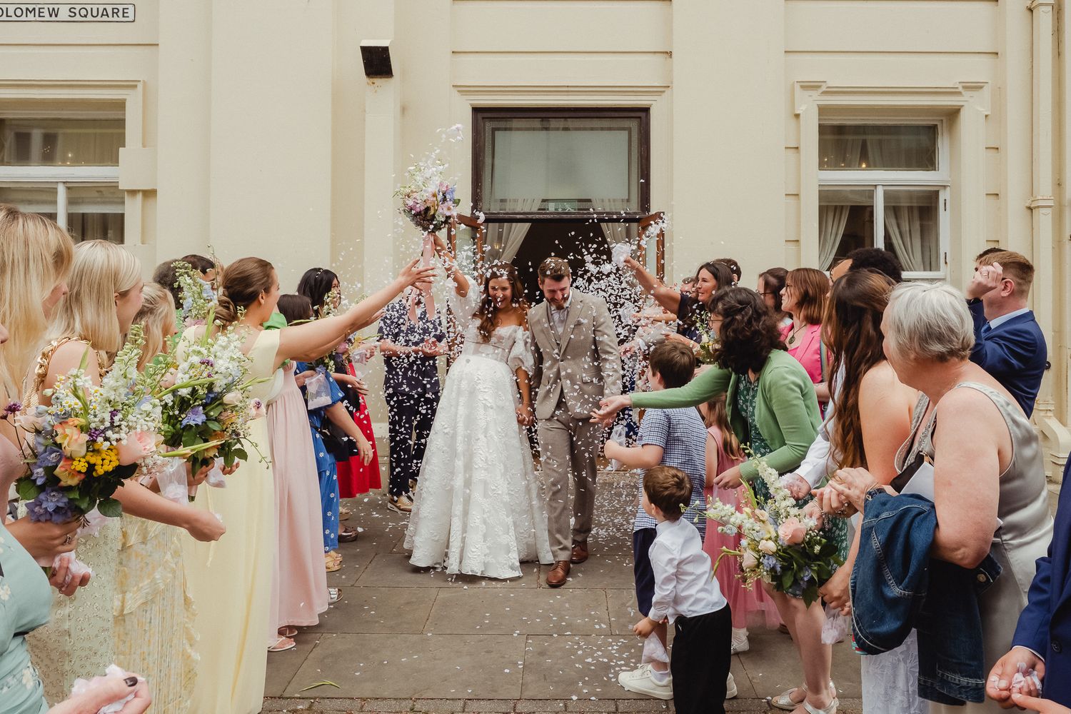 Wedding guests gather outside a historic building for a celebratory confetti toss during a summer ceremony.