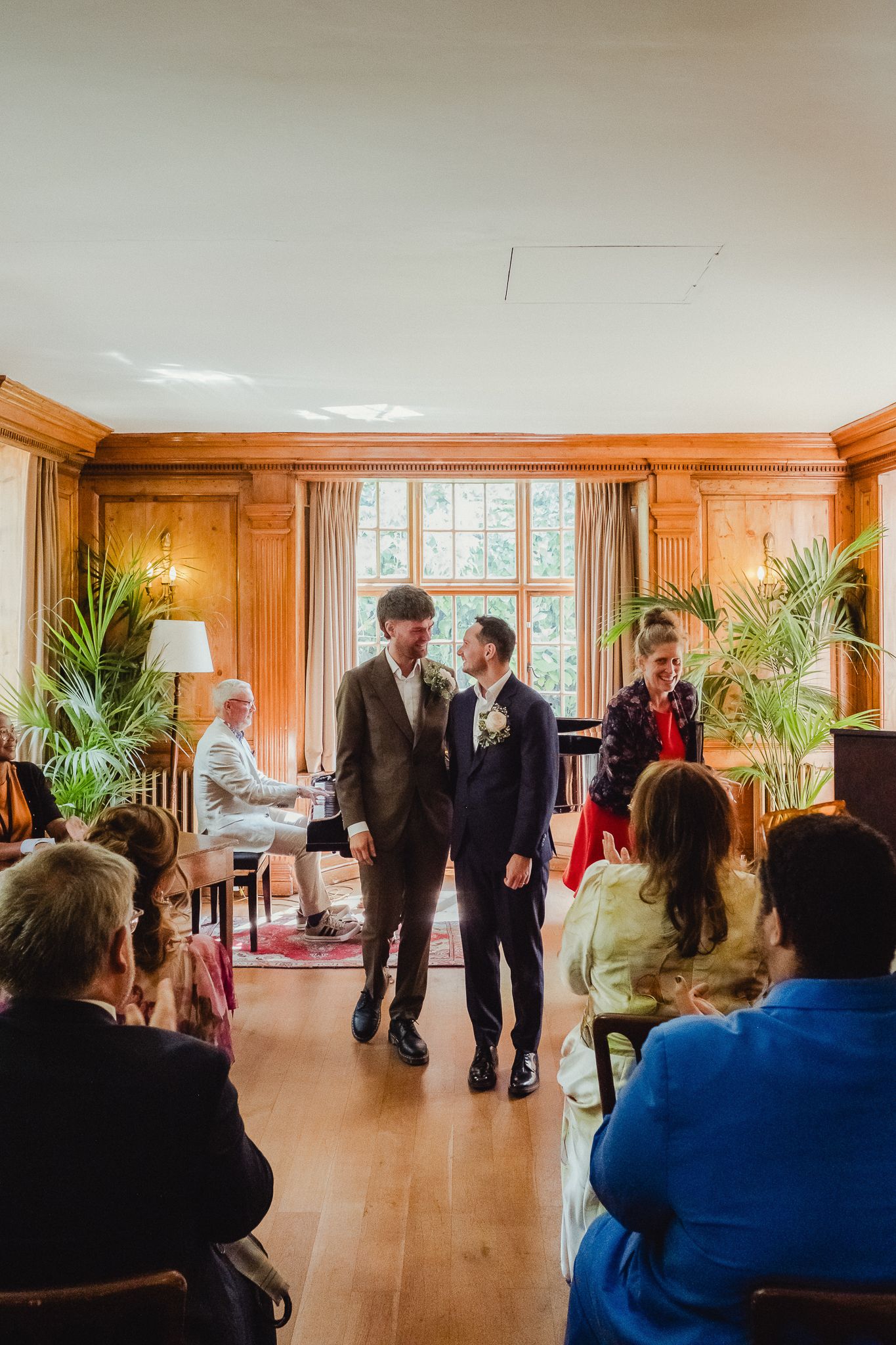 People gather in a warm wood-paneled room with large windows and potted plants for a social event.
