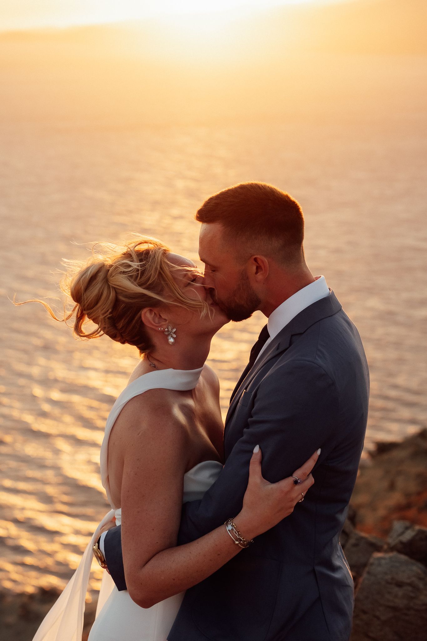 A bride and groom share a romantic kiss at sunset overlooking the ocean on their wedding day.