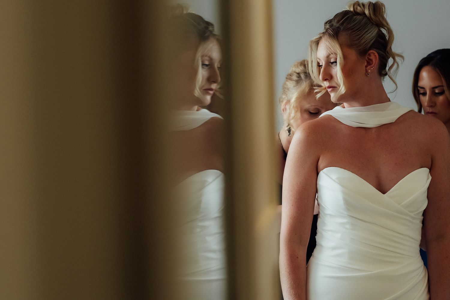 Bride in white strapless wedding gown getting ready, reflected in mirror with bridesmaid assisting.