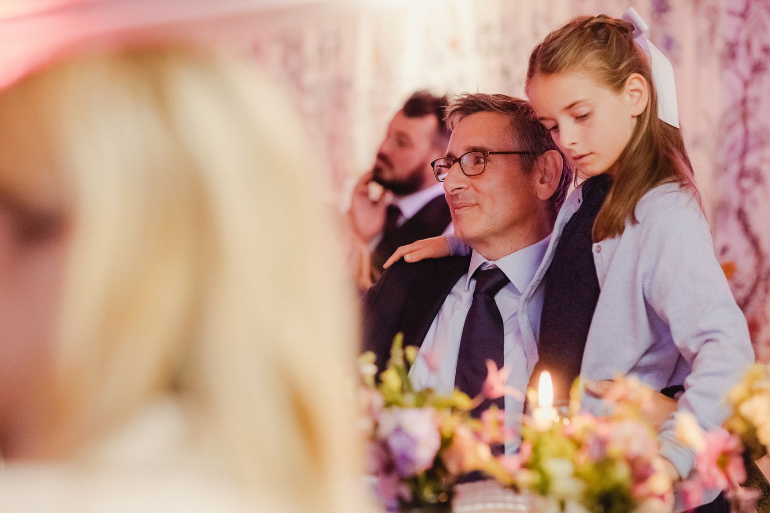 Wedding guests seated at a long reception table decorated with floral centerpieces enjoy conversation and dining.