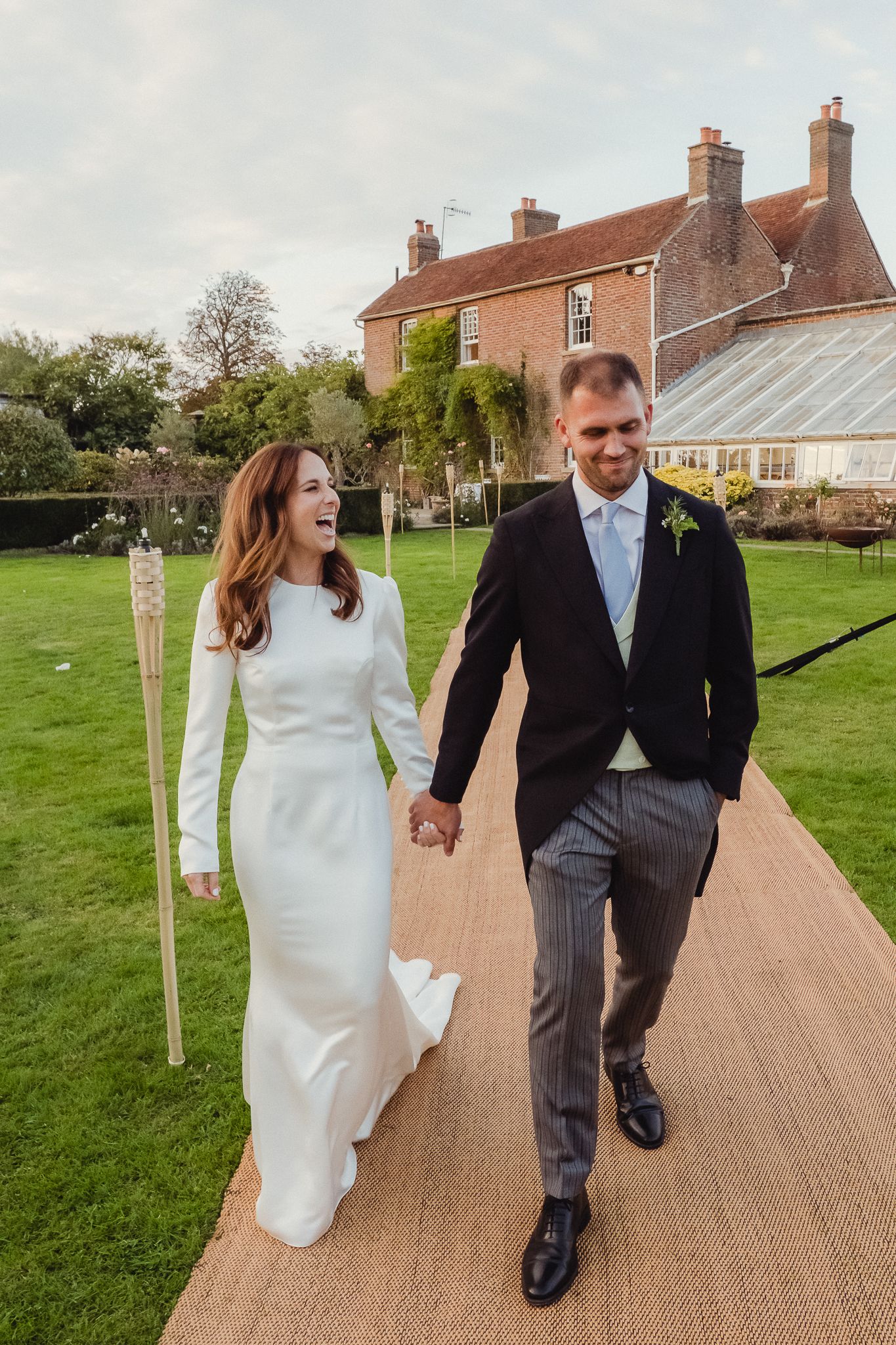 A couple in wedding attire walks together on a brick path through a green lawn on their special day.