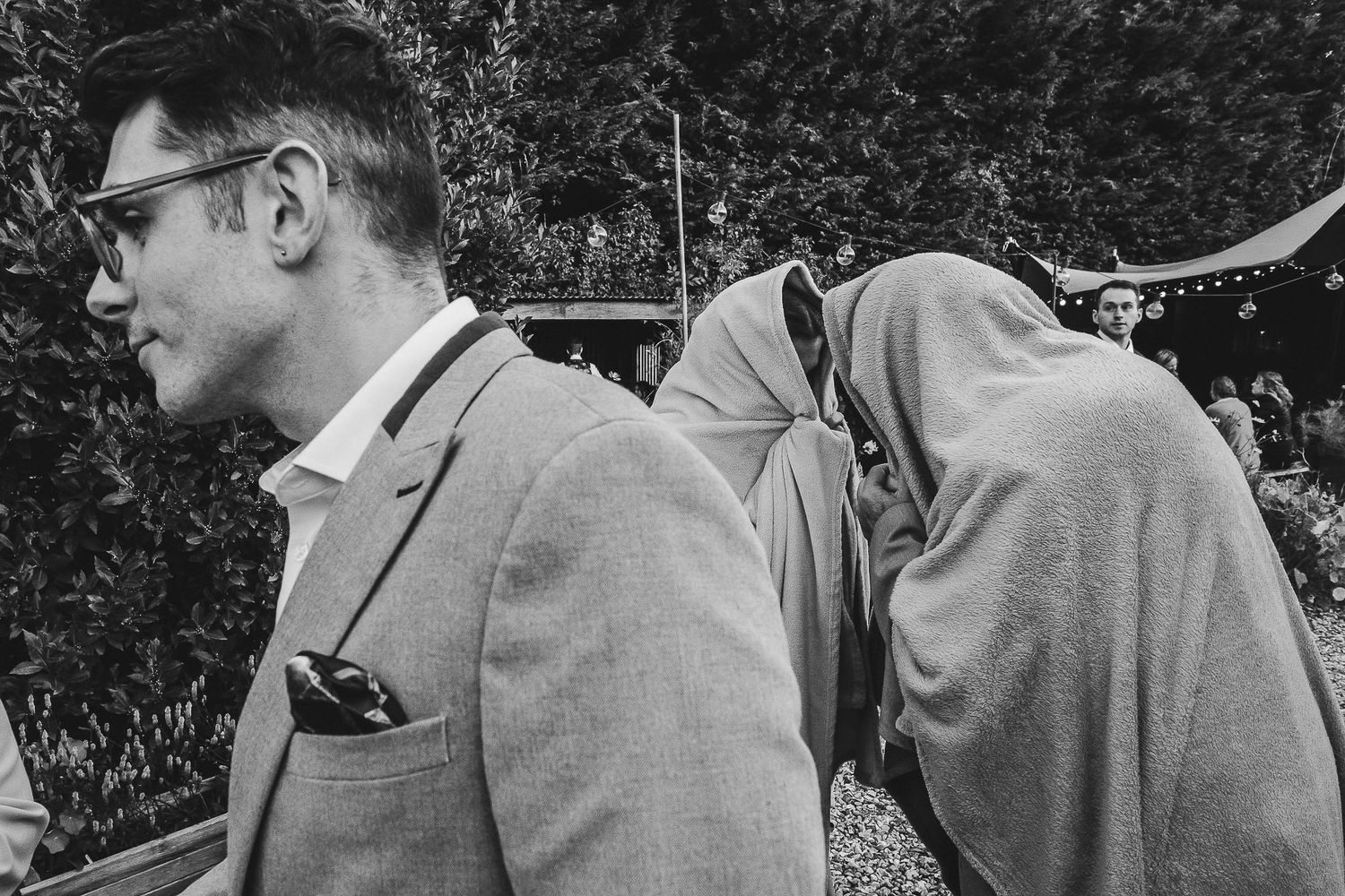 A black and white photograph shows people in formal attire gathered outdoors for a social event.