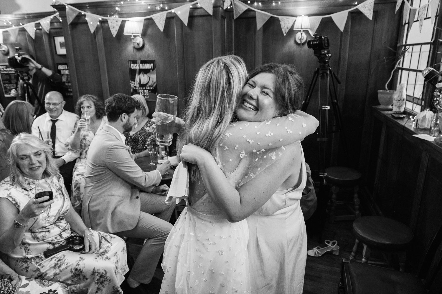 Two women in light dresses share a joyful hug at a wedding reception while seated guests smile and watch in a cozy wood-paneled venue.