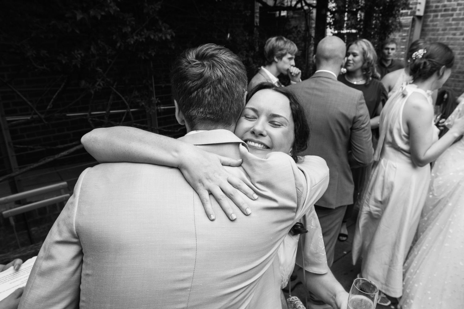 A smiling woman embraces a man in a light suit during a joyful black and white wedding celebration as guests mingle behind them.