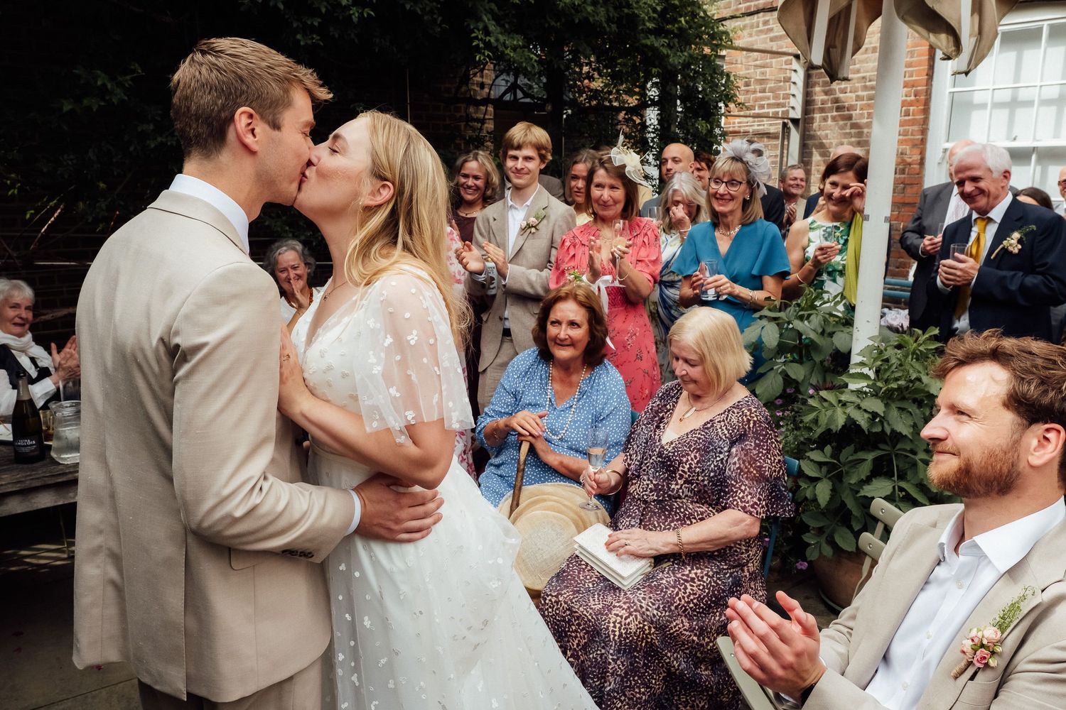 Bride and groom share a kiss during an outdoor wedding ceremony as smiling guests look on and applaud.