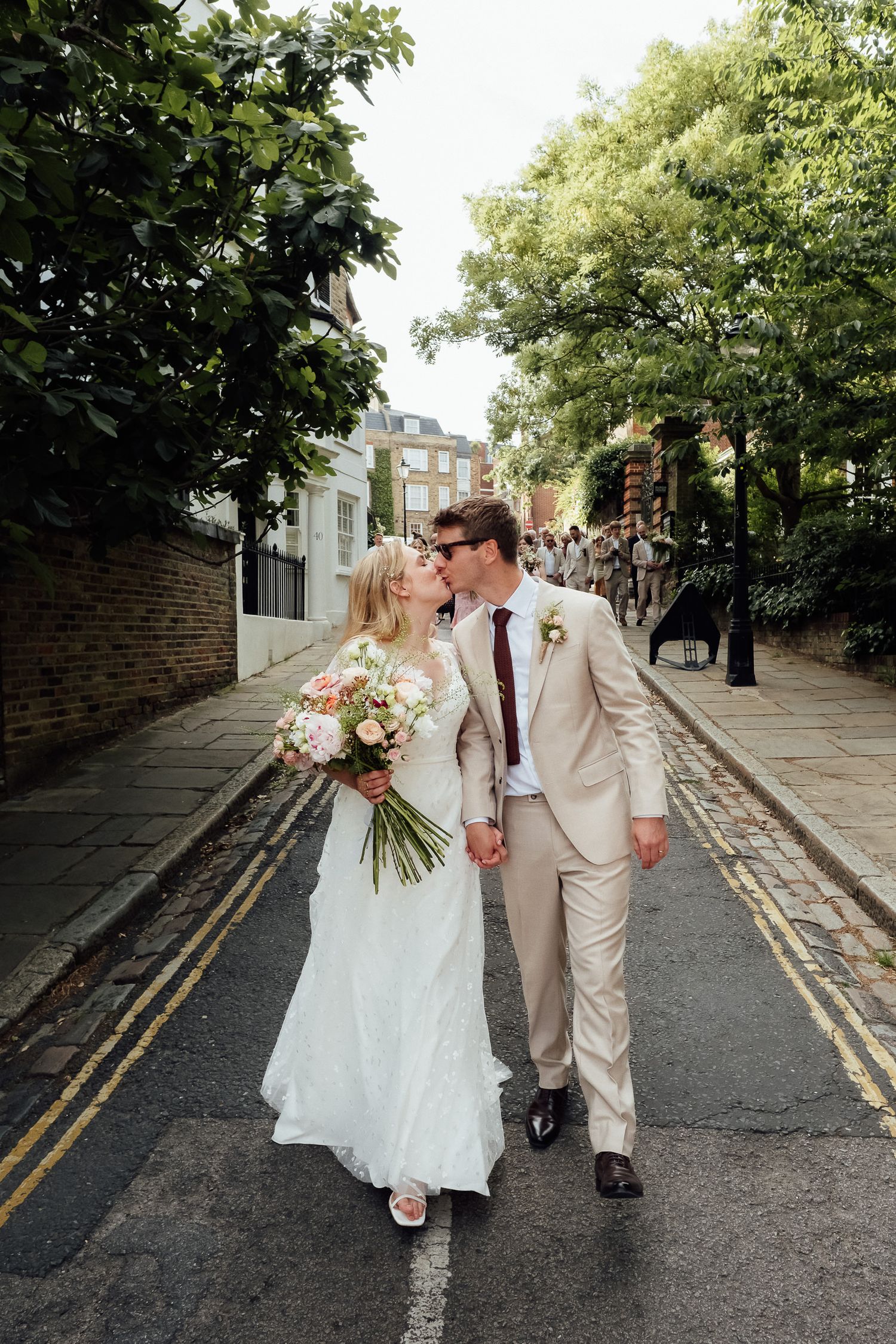 Bride in a white gown holding a pastel bouquet kisses her groom in a beige suit while walking down a quiet tree-lined street.