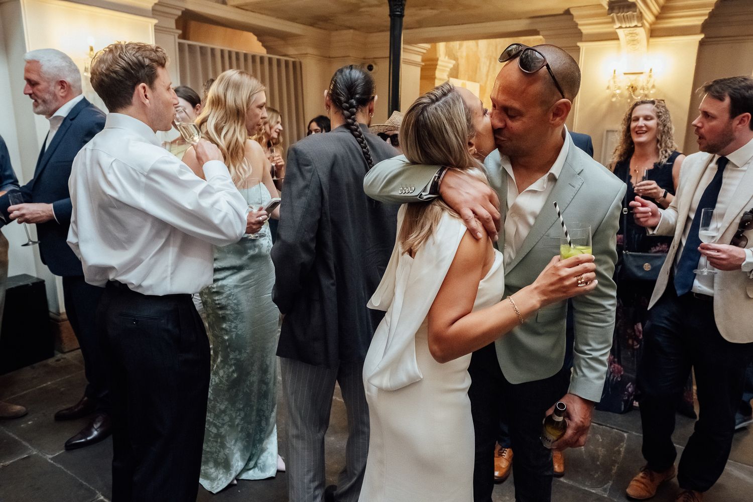 Bride in a white dress embraces a smiling man in a gray suit while wedding guests mingle and talk around them at an indoor reception.