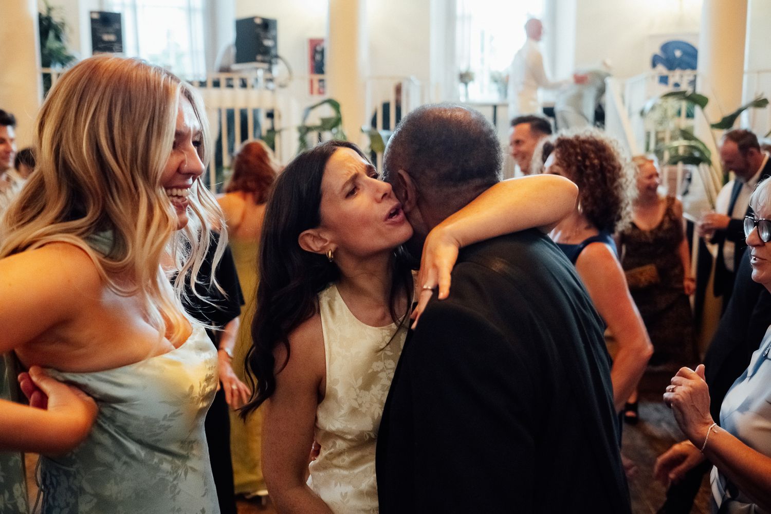 Guests embrace and smile warmly during a lively indoor celebration in a bright, elegant venue with large windows and plants.