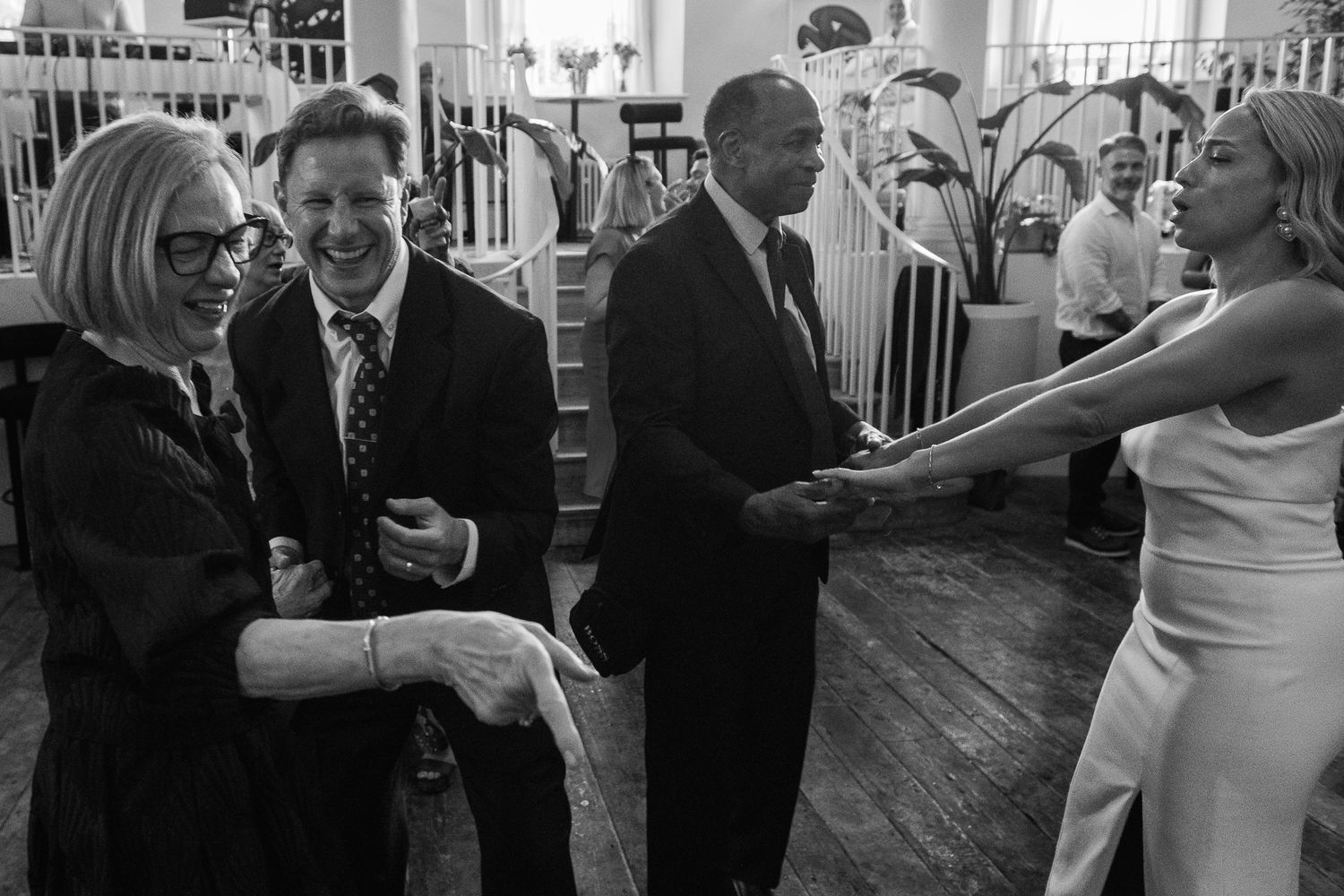 Black and white photo of guests dancing and laughing together on a wooden dance floor at an indoor wedding reception.