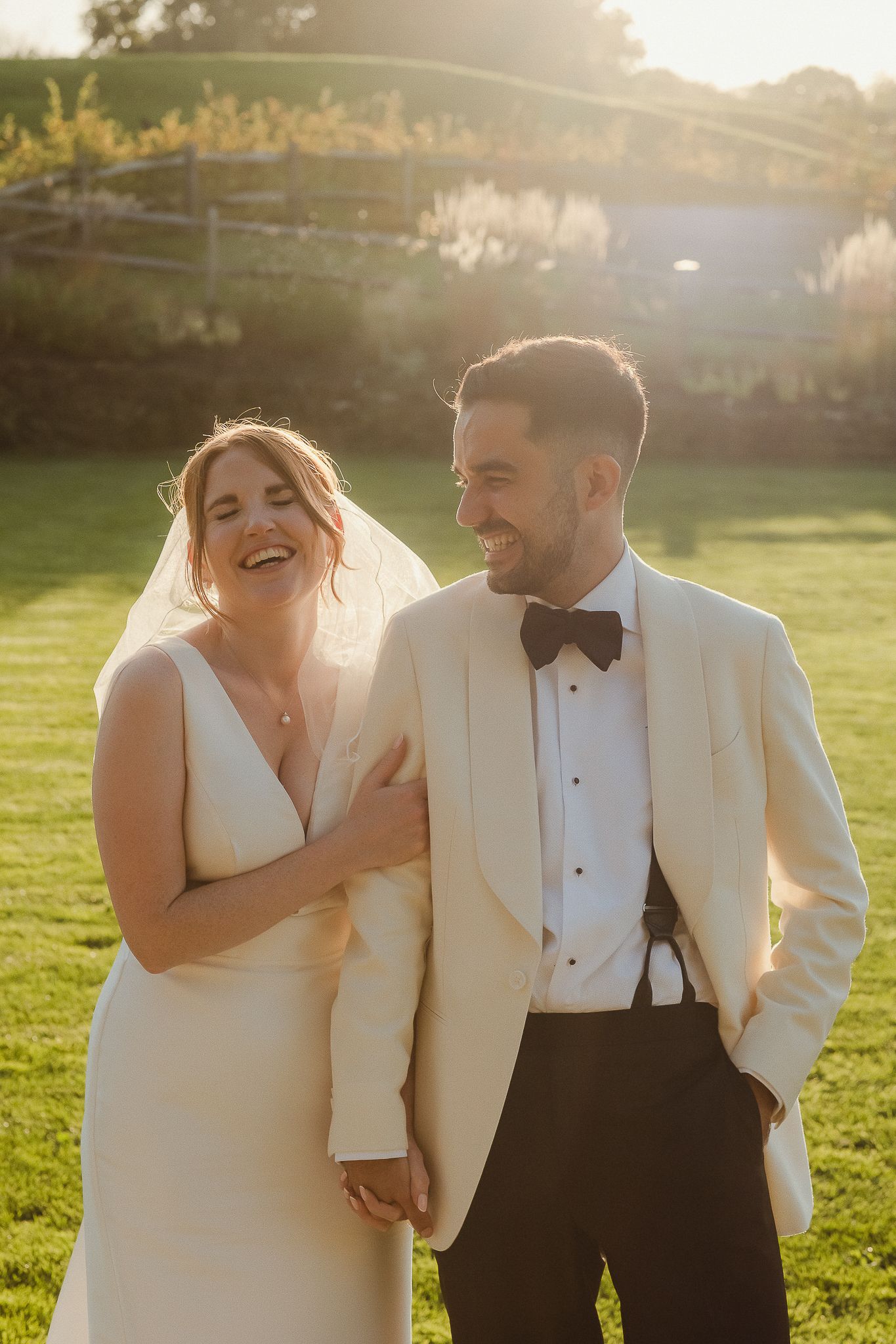 A joyful couple shares candid moments in a sunlit field wearing formal white and black attire during their wedding photoshoot.