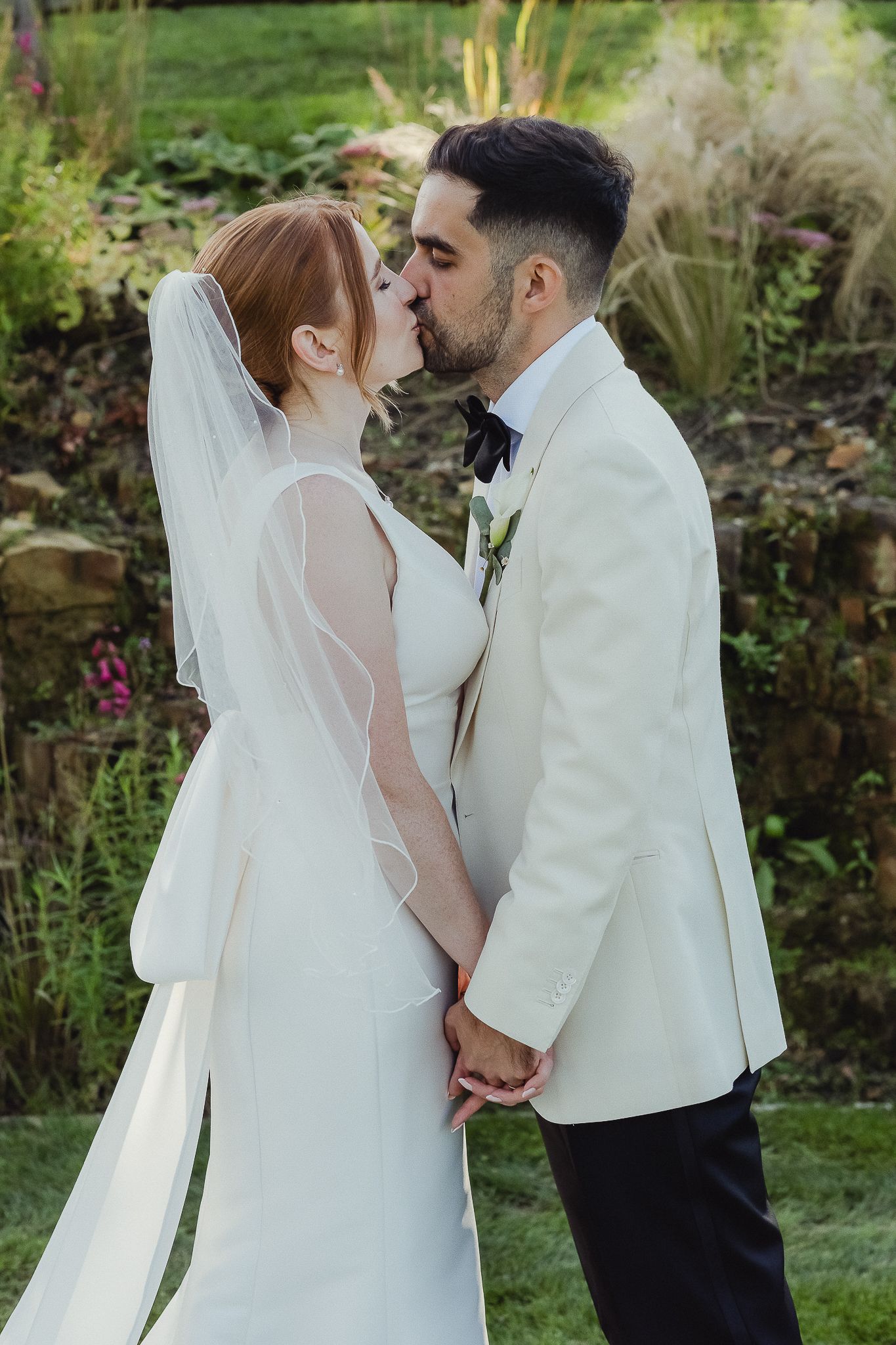 A romantic wedding kiss between newlyweds in white attire surrounded by a lush garden setting.