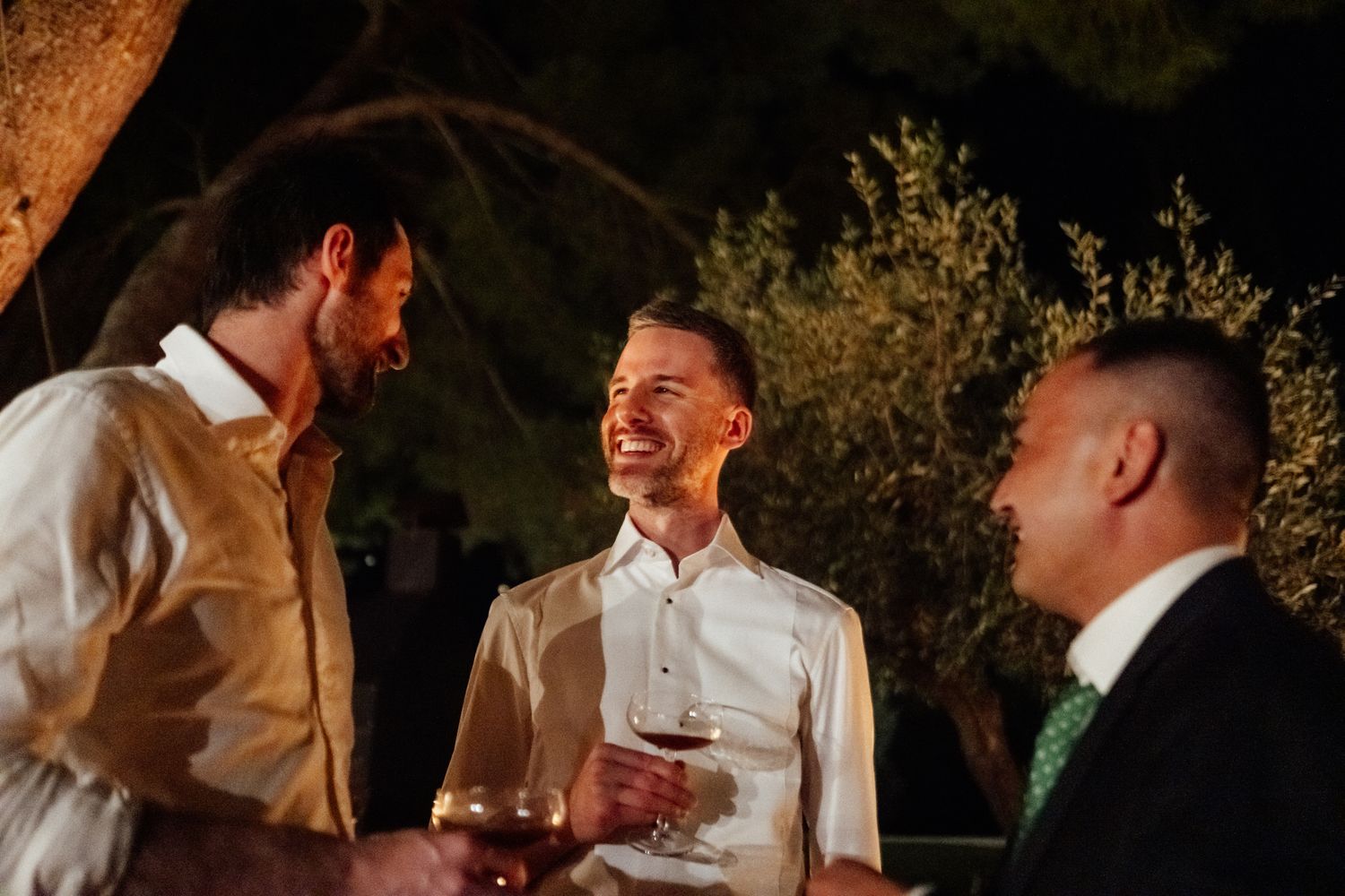 Three men in light dress shirts chat and laugh while holding wine glasses at an evening outdoor gathering under olive trees.