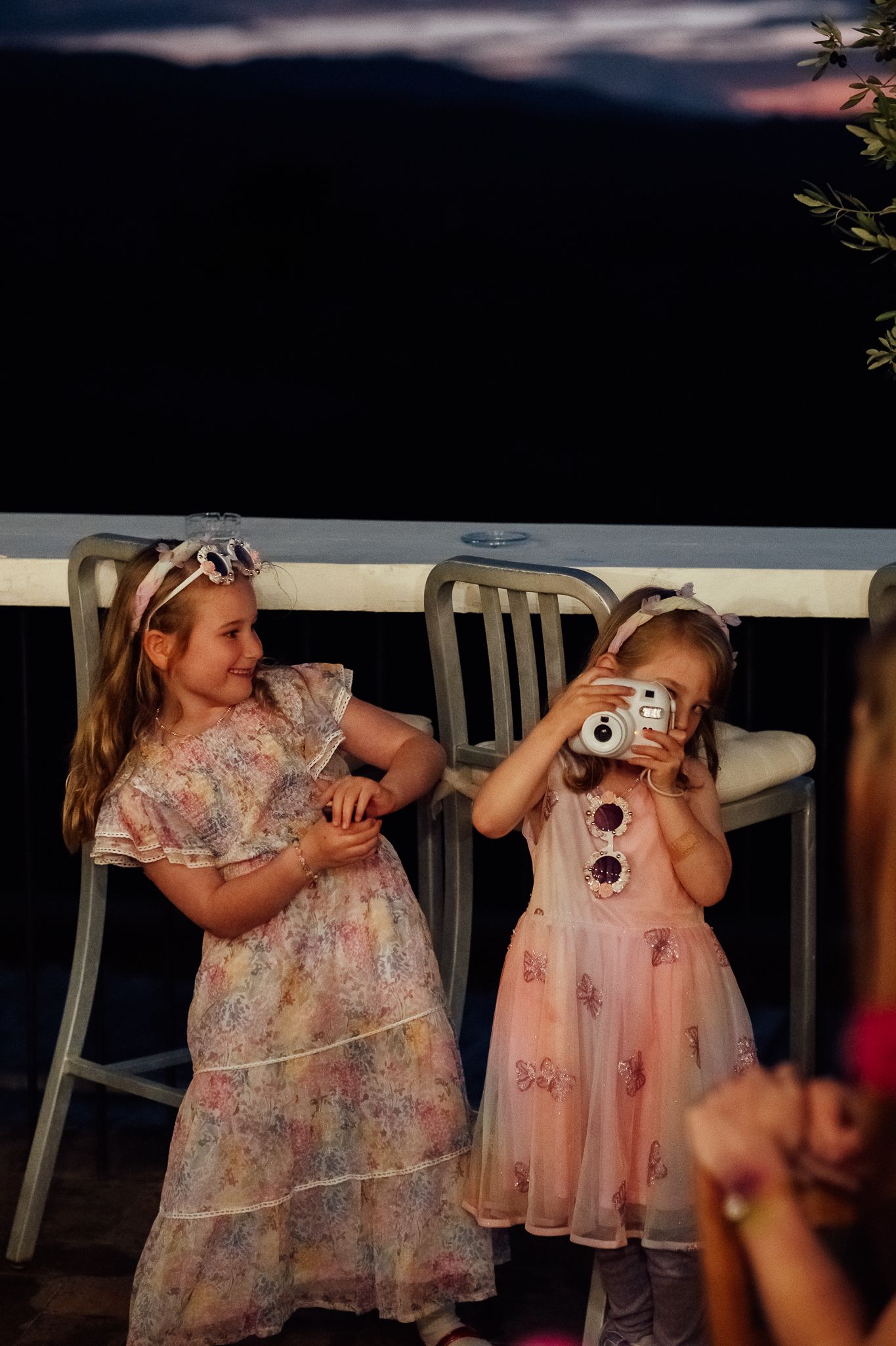 Two young girls in floral dresses and headbands play on an outdoor terrace at dusk, one taking a photo with a small instant camera.