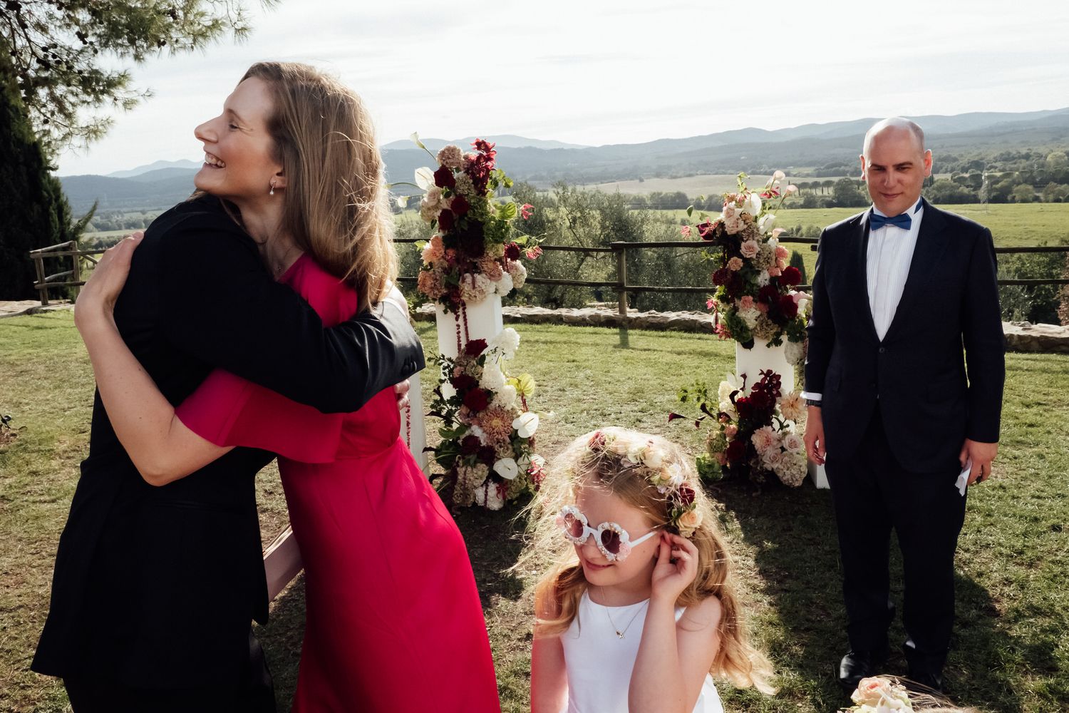 Woman in a red dress hugs a guest at an outdoor wedding while a girl in heart sunglasses and a man in a suit stand near floral arrangements.