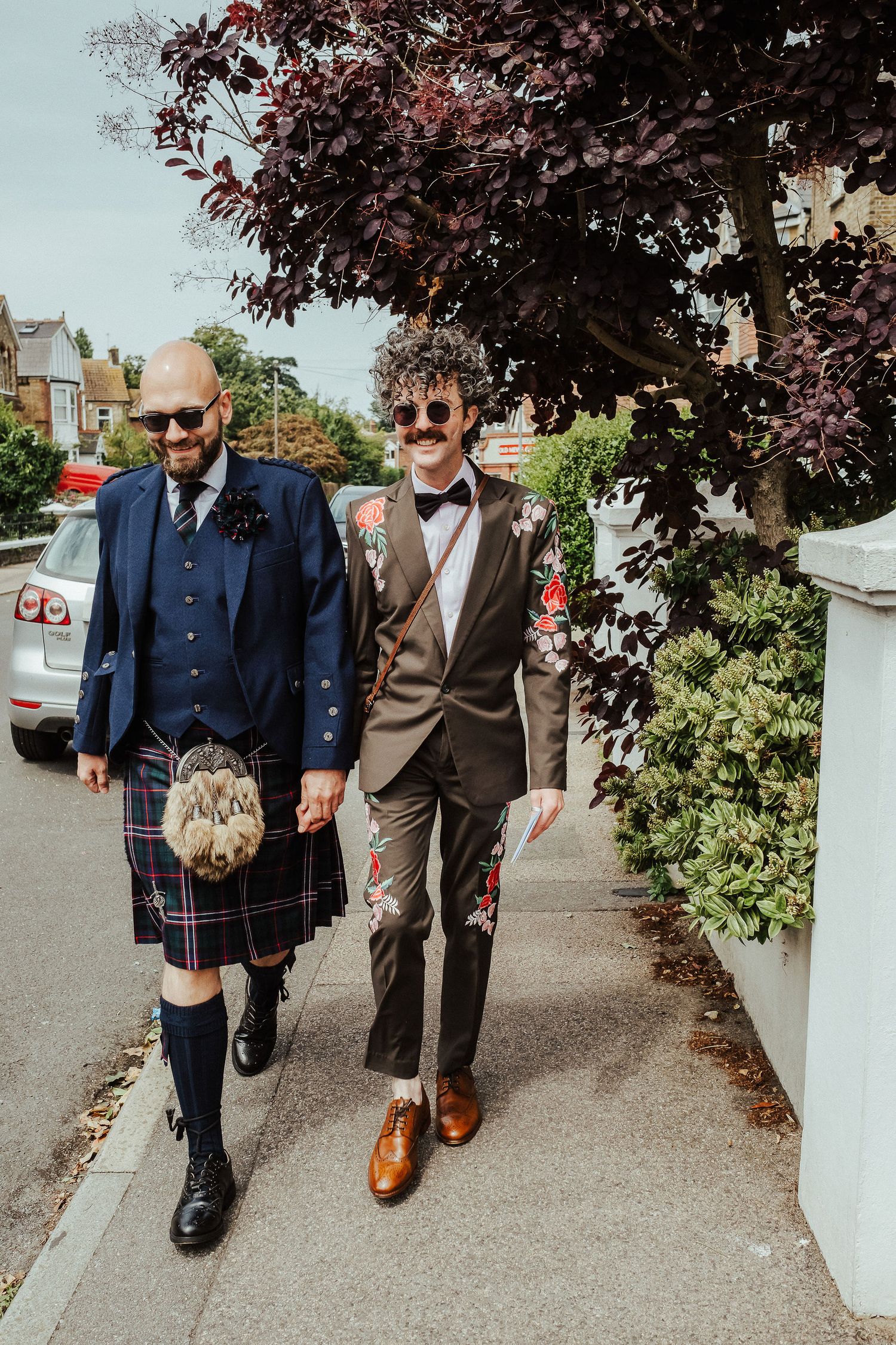 Two men in wedding attire hold hands and walk down a tree-lined sidewalk, one in a navy jacket and tartan kilt, the other in a floral-embroi