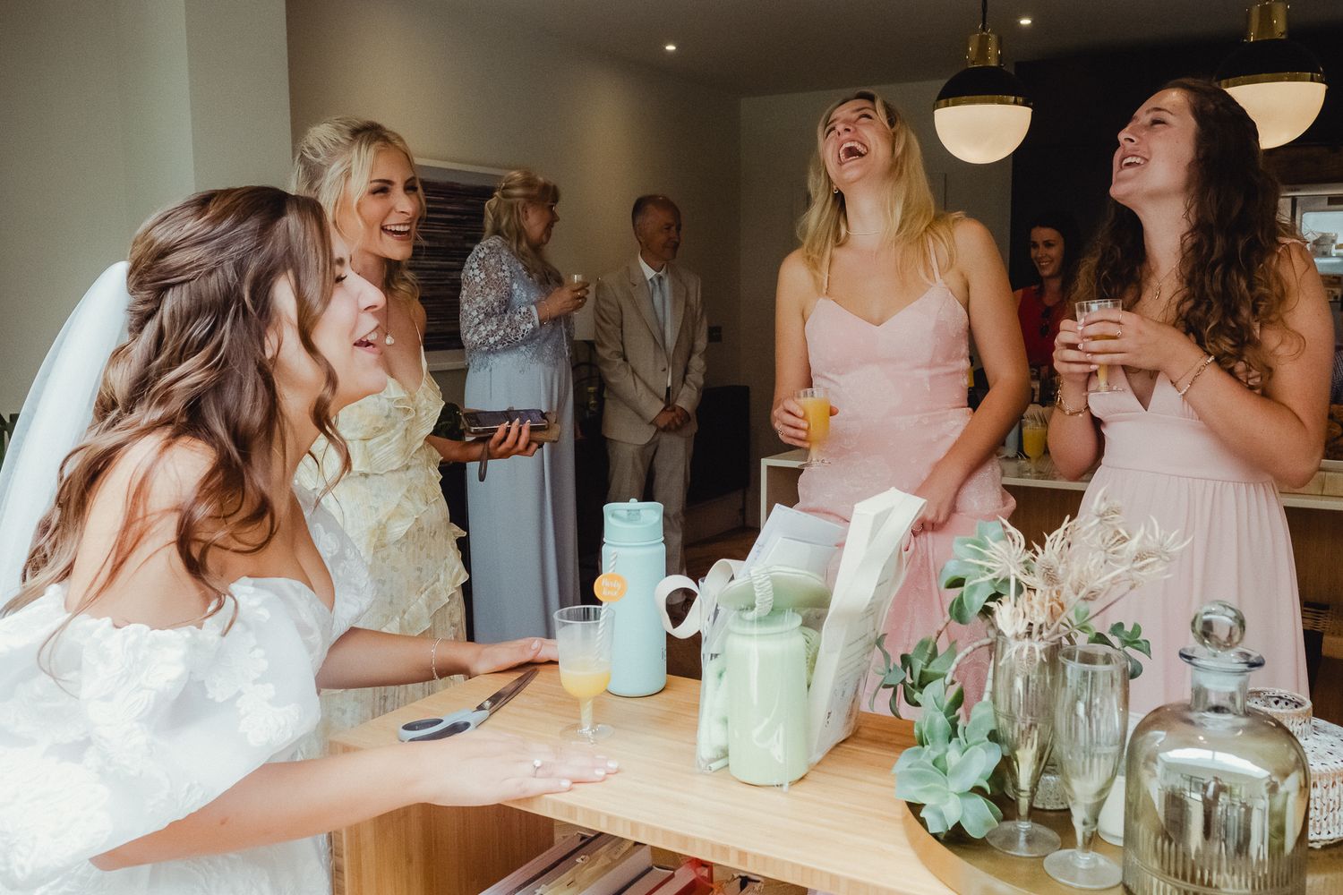 Wedding party guests gather around a table enjoying drinks and laughter at an elegant indoor reception.
