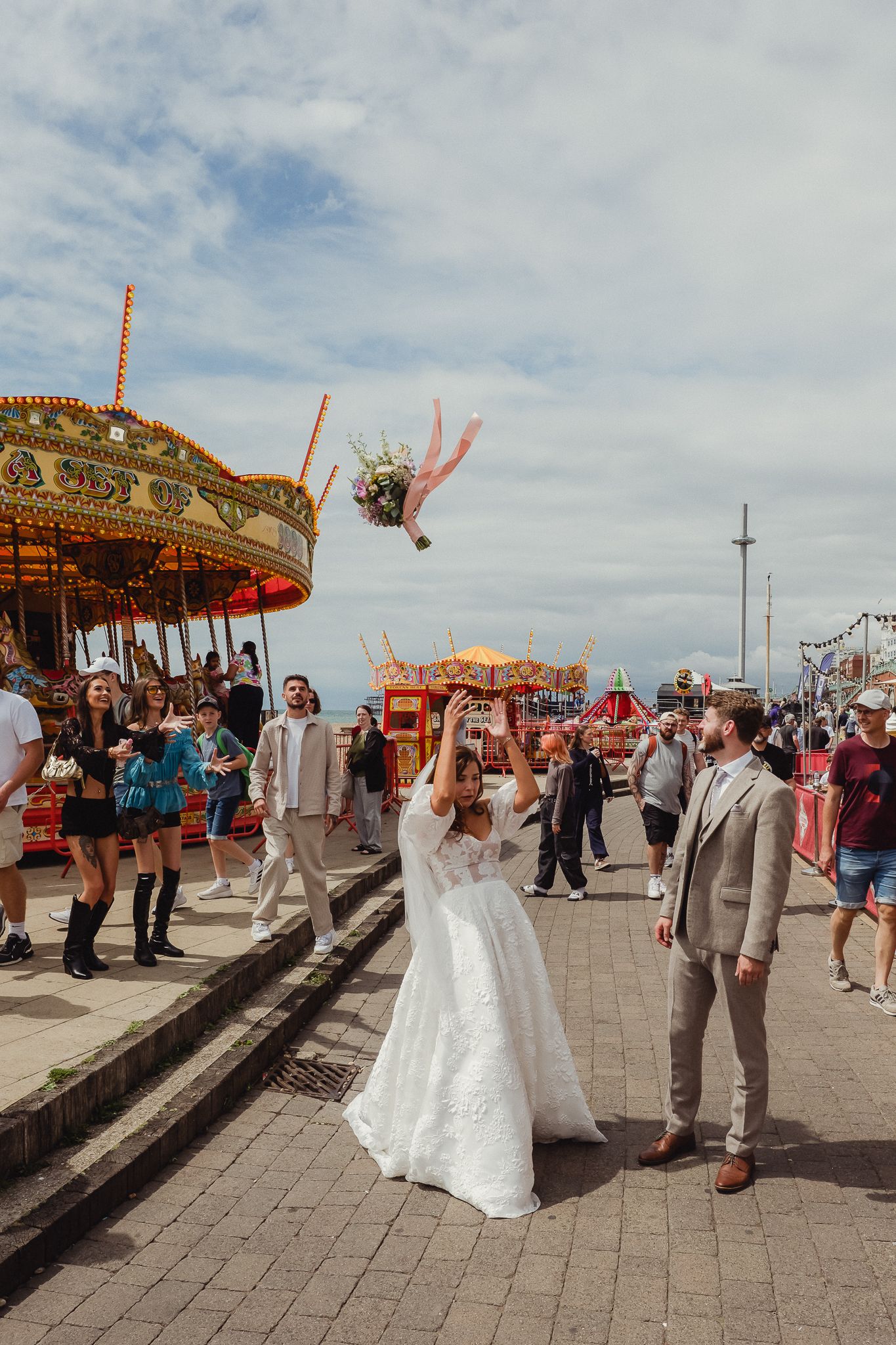 A couple celebrates their wedding day at a carnival with a vintage carousel in the background on a cloudy afternoon.