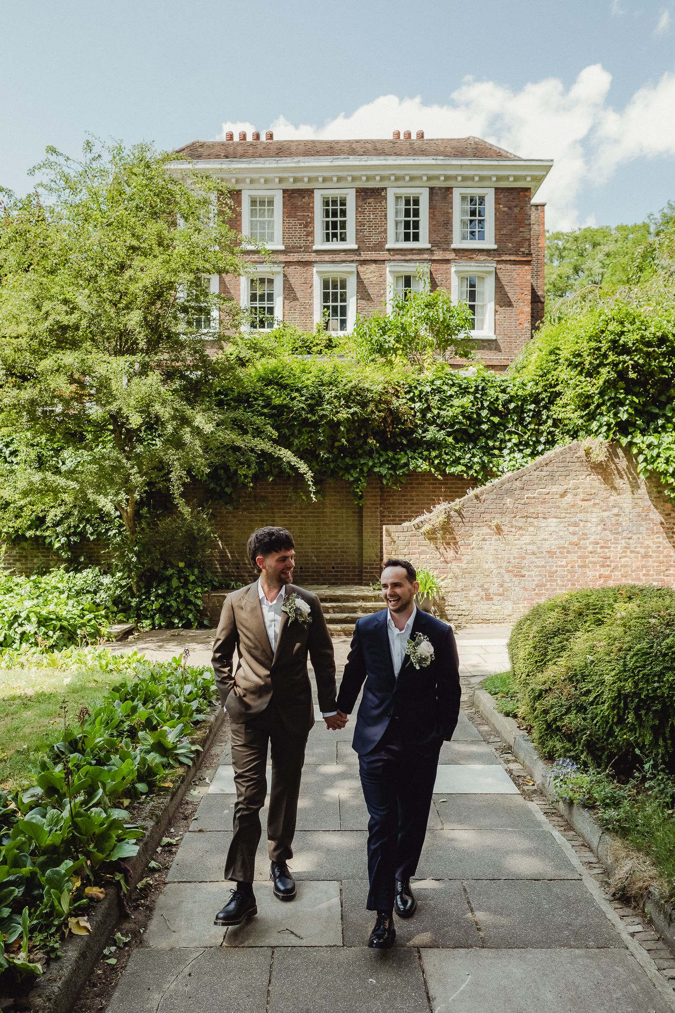 Wedding party walks along a garden path beside a brick wall at an elegant Georgian manor house on a sunny day.