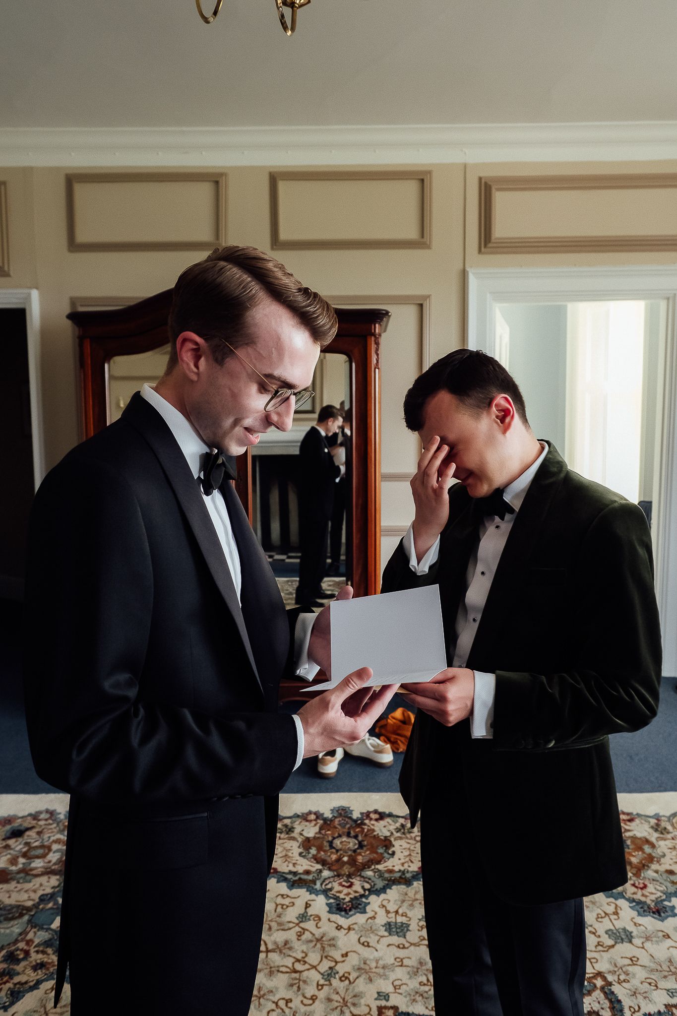 Two grooms in tuxedos share an emotional moment reading wedding vows in an elegant room before their ceremony.