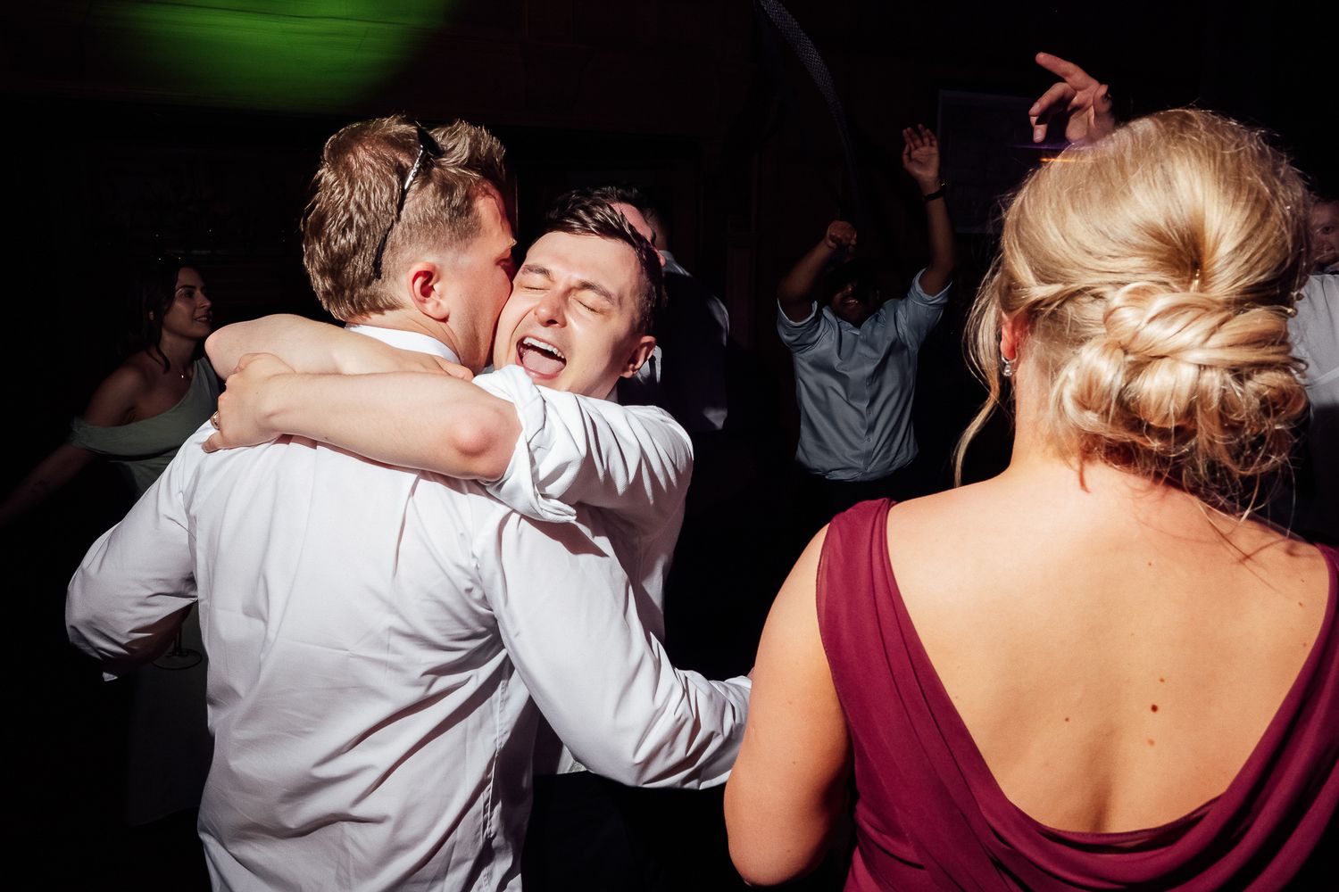 Two men hugging and dancing joyfully at a wedding reception party with a woman in burgundy dress nearby.