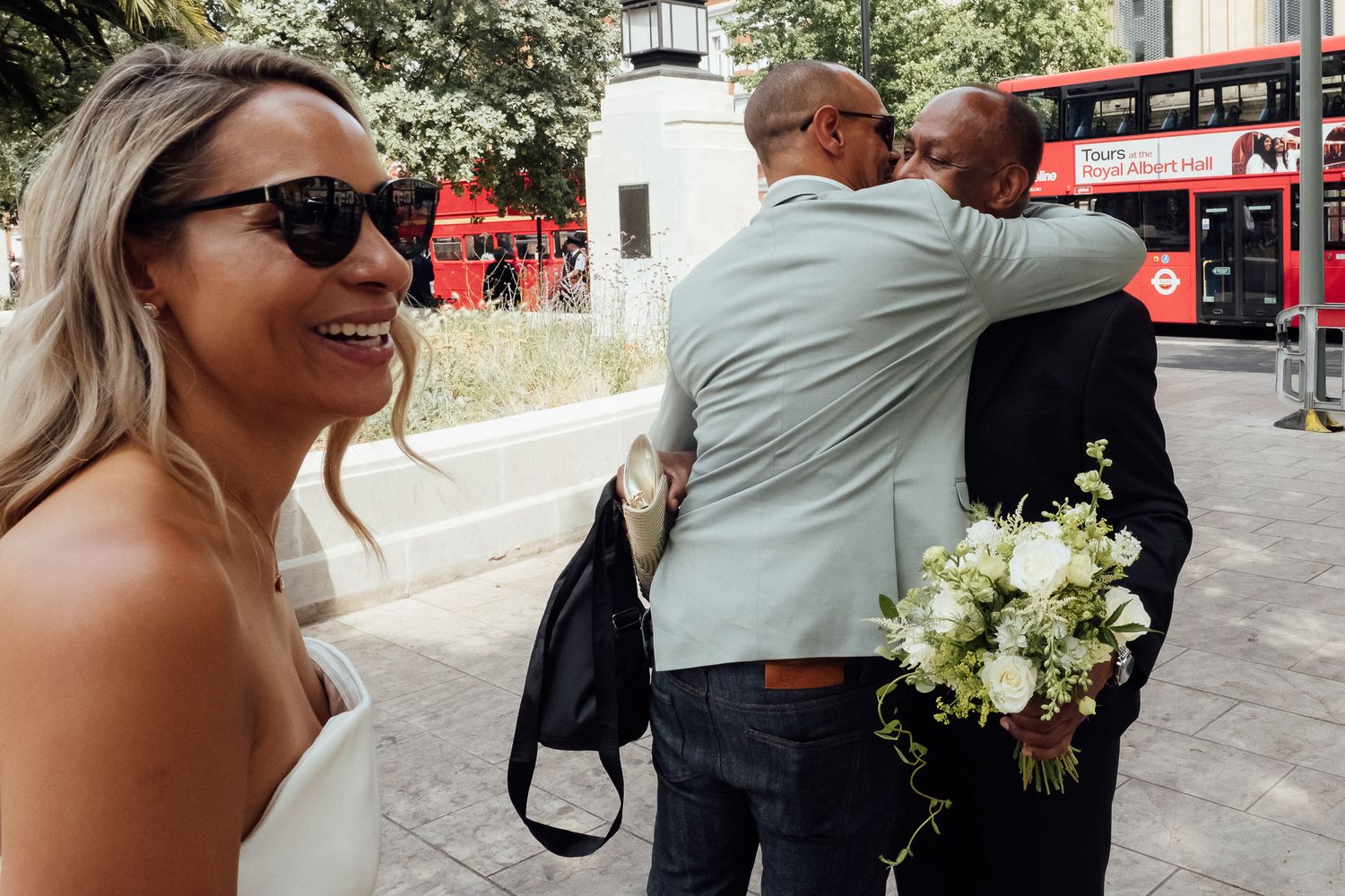 Smiling woman in sunglasses and a white dress holds a bridal bouquet as two men embrace on a city street near a red London bus.