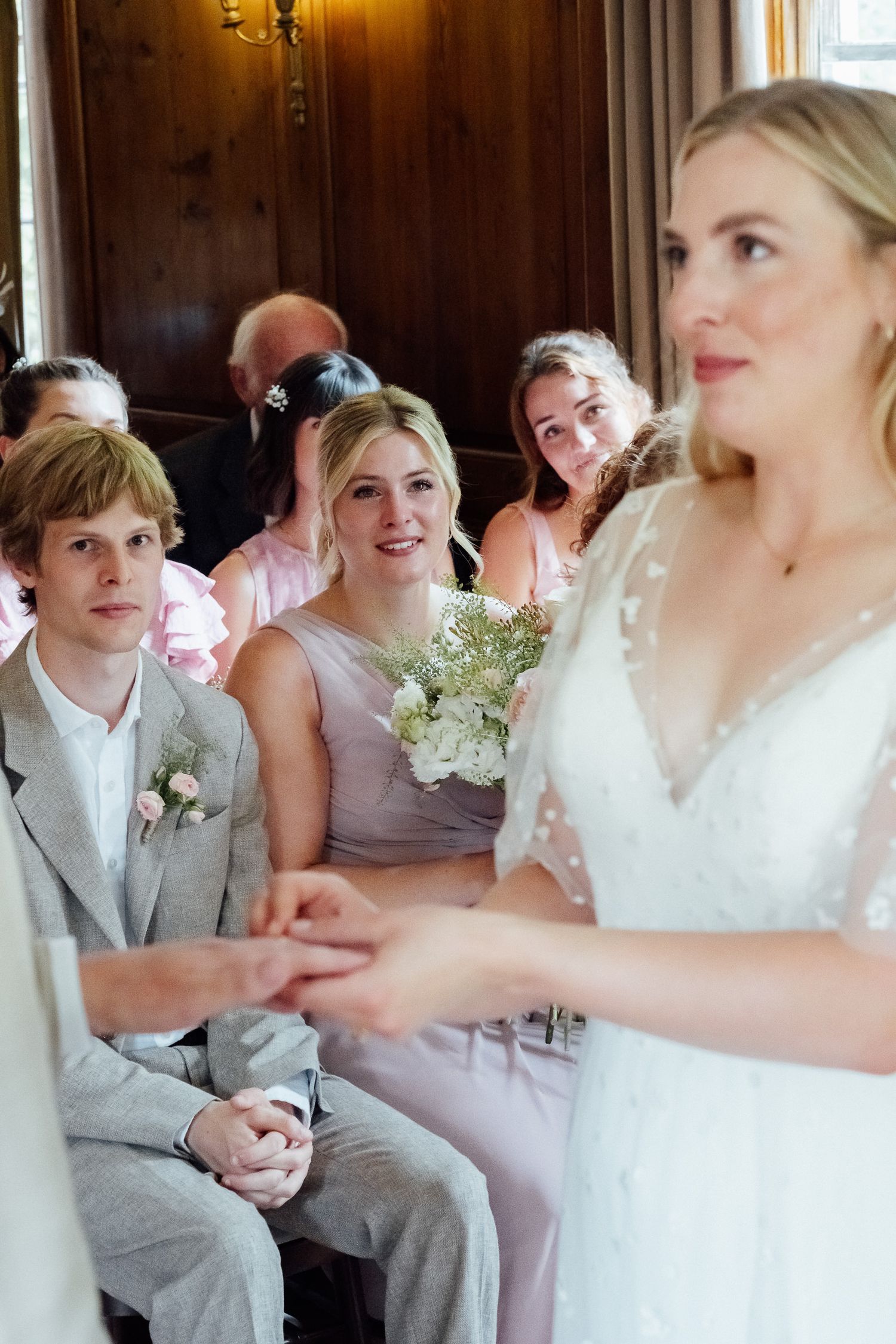 Bride in a white lace gown exchanges vows and holds hands with her partner as smiling wedding guests watch from wooden pews.