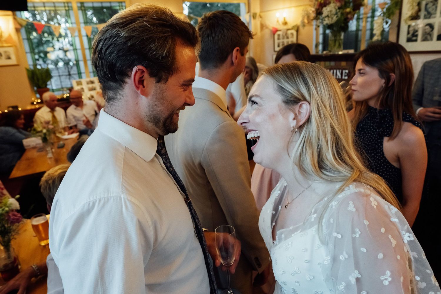 Smiling bride in a white lace dress laughs with a groom in a white shirt and tie at a lively indoor wedding reception with guests.