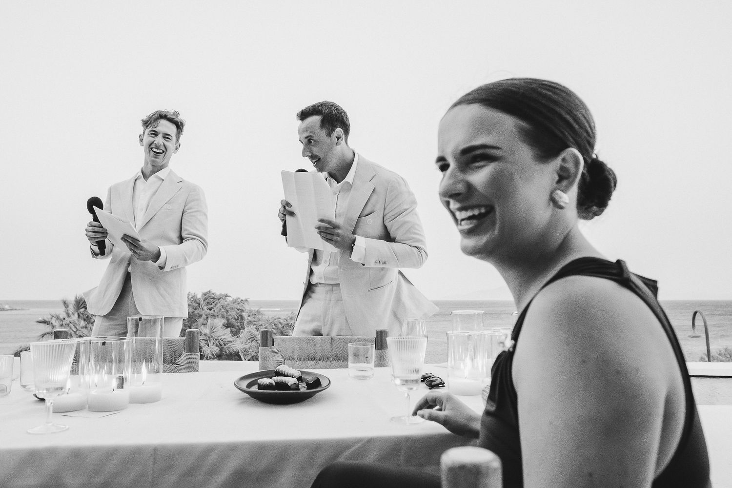 A group enjoys conversation and food around a sunny outdoor dining table with plates and dishes spread out.