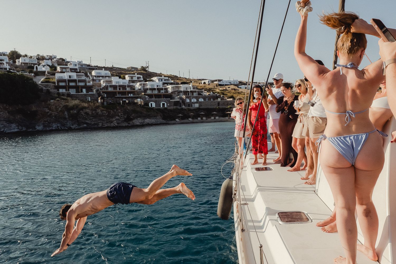 People enjoying a summer boat party with swimming and diving off a yacht near a coastal Mediterranean town.