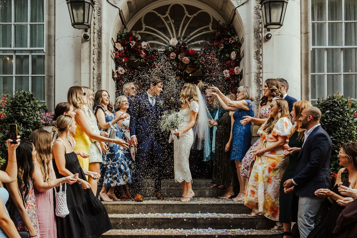 A joyful wedding celebration scene with guests gathering on stone steps outside a historic building entrance.