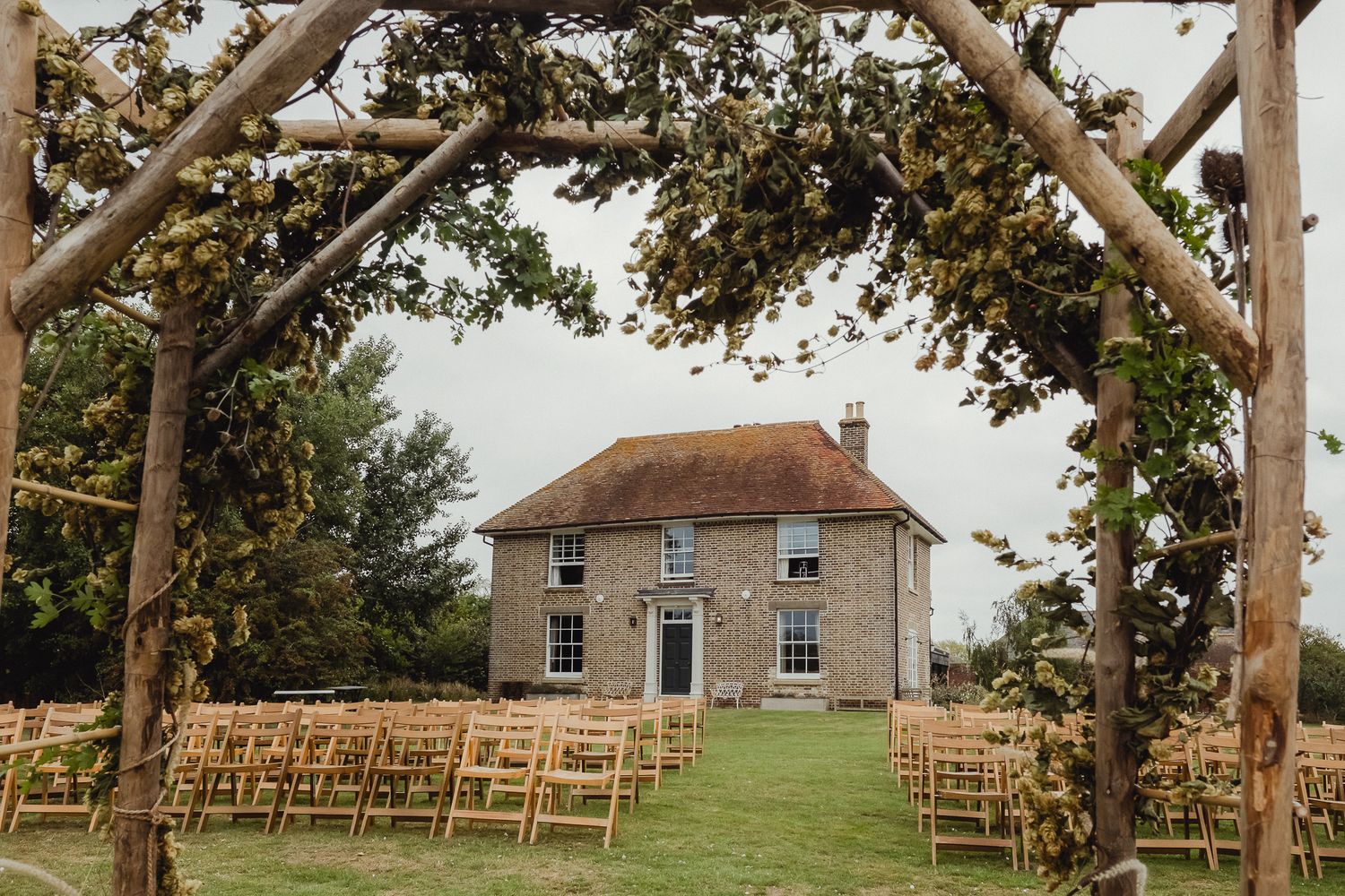 Rustic outdoor wedding ceremony setup with wooden chairs arranged under tall trees beside a historic stone house.