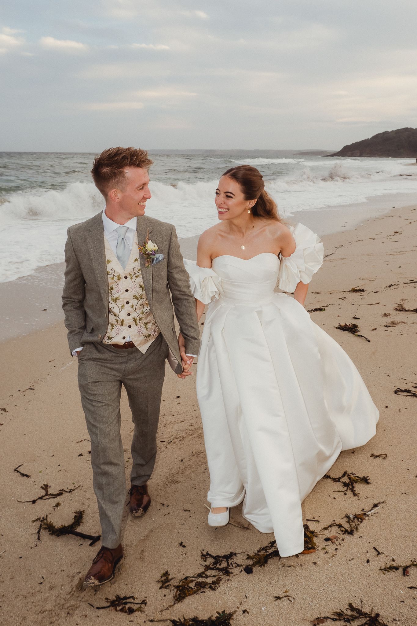 A couple walks hand in hand along a scenic beach shoreline wearing wedding attire on an overcast day.
